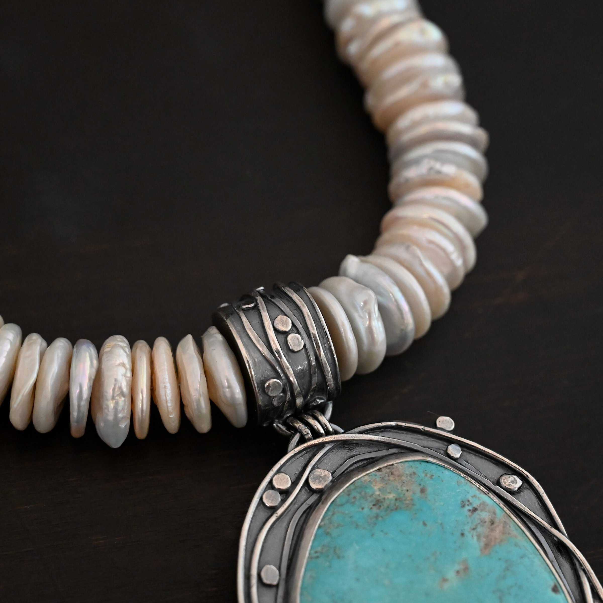 Calypso Necklace Photo of the "Calypso" Necklace. It is a sterling silver, turquoise, and pearl necklace. There is a large, oval shaped turquoise. It is set in sterling silver. The inside of the silver setting has hammered silver dots going all around it, and the outside of the silver setting has swirling silver lines going all around the turquoise. The collar of the necklace is made entirely out of strung pearl disks that are large and rough circle shapes. There is a large silver attachment on the top of the turquoise pendant to attach it to the collar. The attachment has a hole going through the center, and it is in the center of the necklace, with pearls on each side of it. Necklace is laying against a dark background.