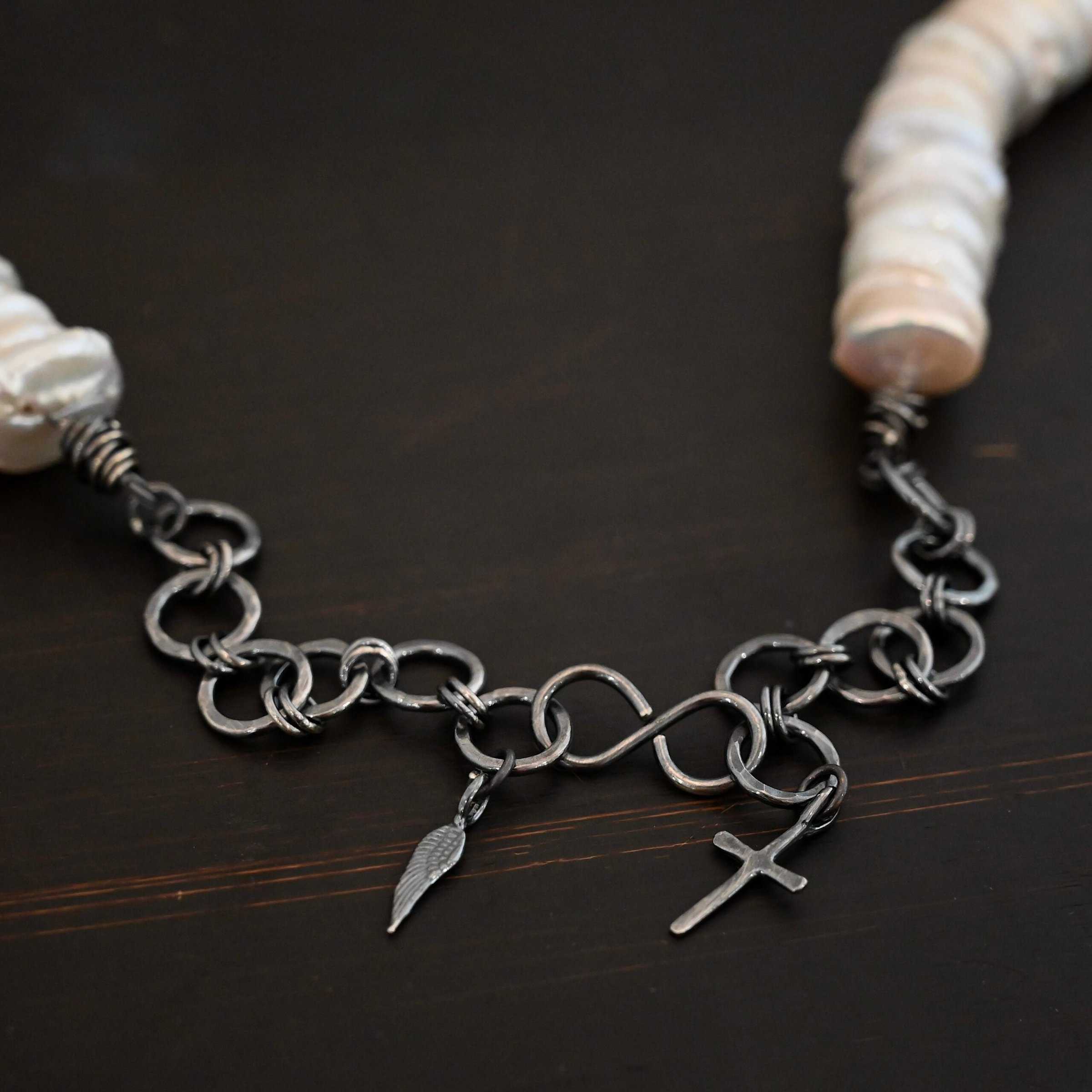Pearl Necklace ends Photo of the "Cho Oyu" Necklace clasp. The collar of the necklace is made entirely out of strung pearl disks that are large and rough circle shapes. The clasp is a thick s hook clasp attached on both sides to hammered circle links that are thick for the extension of the necklace. There is a cross and angel wing charm on both ends of the extension. Clasp is laying against a dark background.
