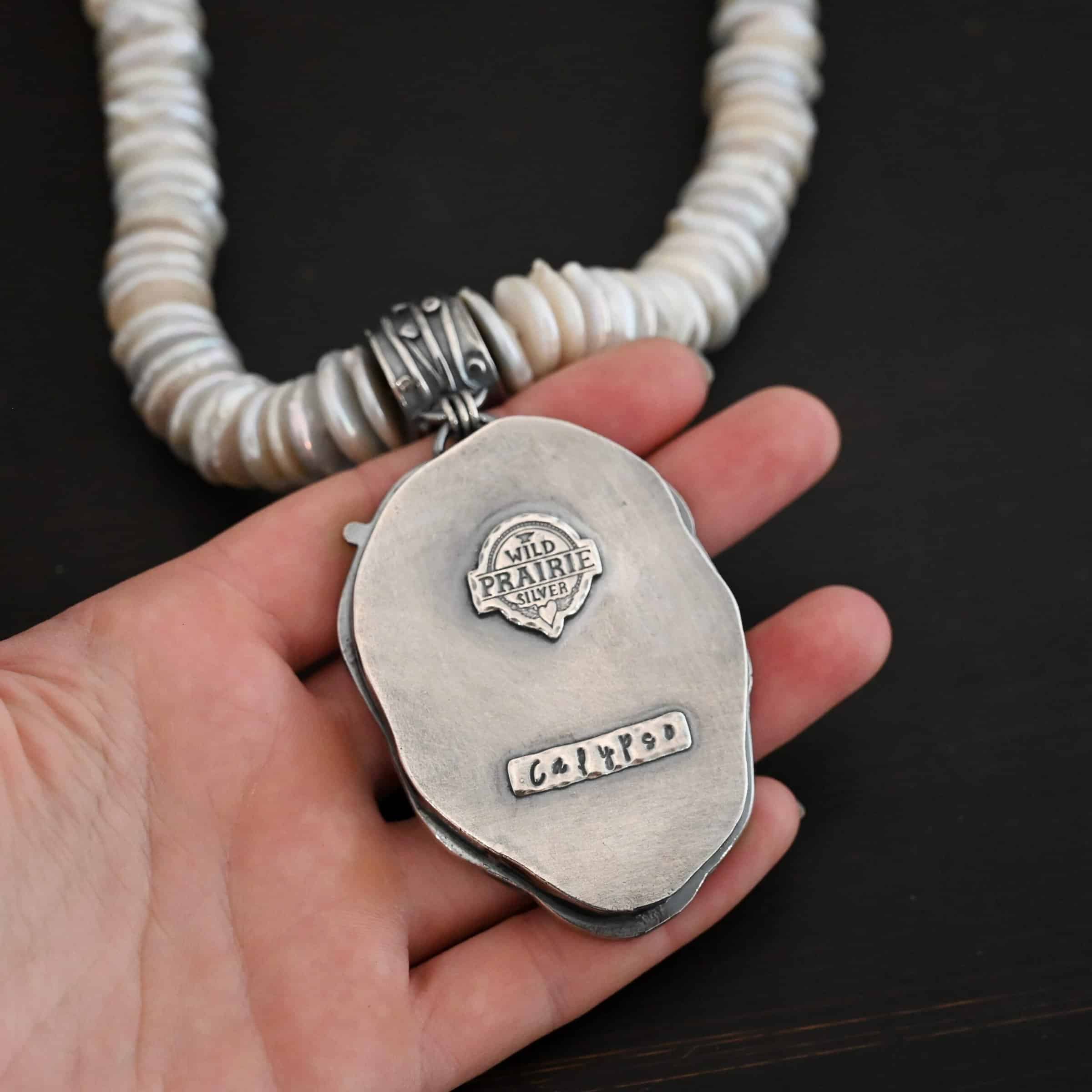 Calypso Necklace Photo of the back of the "Calypso" Necklace. The back of the pendant is entirely sterling silver. The top of it has the "Wild Prairie Silver" Logo soldered to it with a shiny rectangle that has the word "Calypso" stamped into it. The collar of the necklace is made entirely out of strung pearl disks that are large and rough circle shapes. There is a large silver attachment on the top of the turquoise pendant to attach it to the collar. The attachment has a hole going through the center, and it is in the center of the necklace, with pearls on each side of it. Necklace is laying against the palm of a hand to demonstrate size.