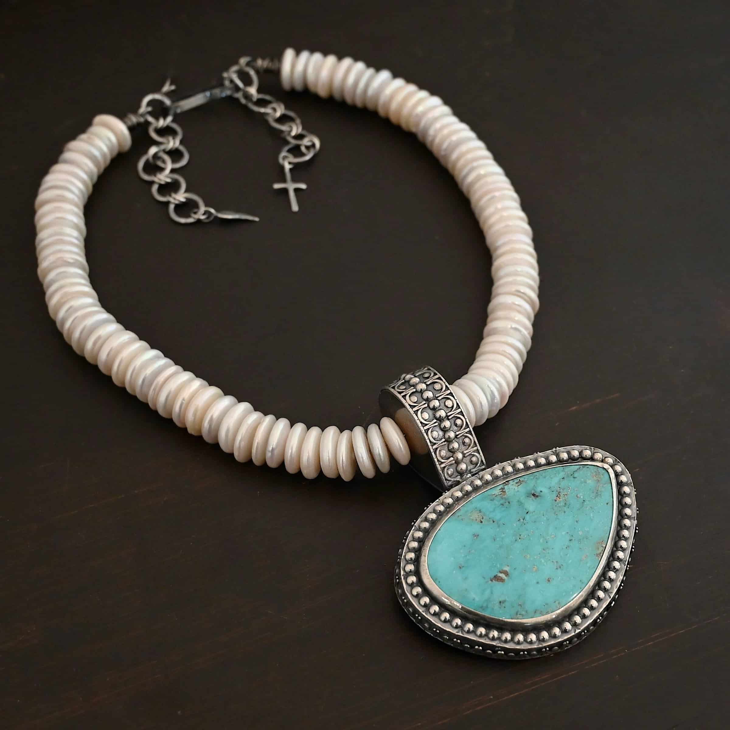 Photo of the "Cho Oyu" Necklace. It is a sterling silver, turquoise, and pearl necklace. There is a large, oval shaped turquoise set horizontally. It is set in sterling silver. The outside of the silver has silver balls set right next to each other going all the way around the turquoise. The collar of the necklace is made entirely out of strung pearl disks that are large and rough circle shapes. There is a large silver attachment on the top of the turquoise pendant to attach it to the collar. The attachment has a hole going through the center, and it is in the center of the necklace, with pearls on each side of it. Necklace is laying against a dark background.