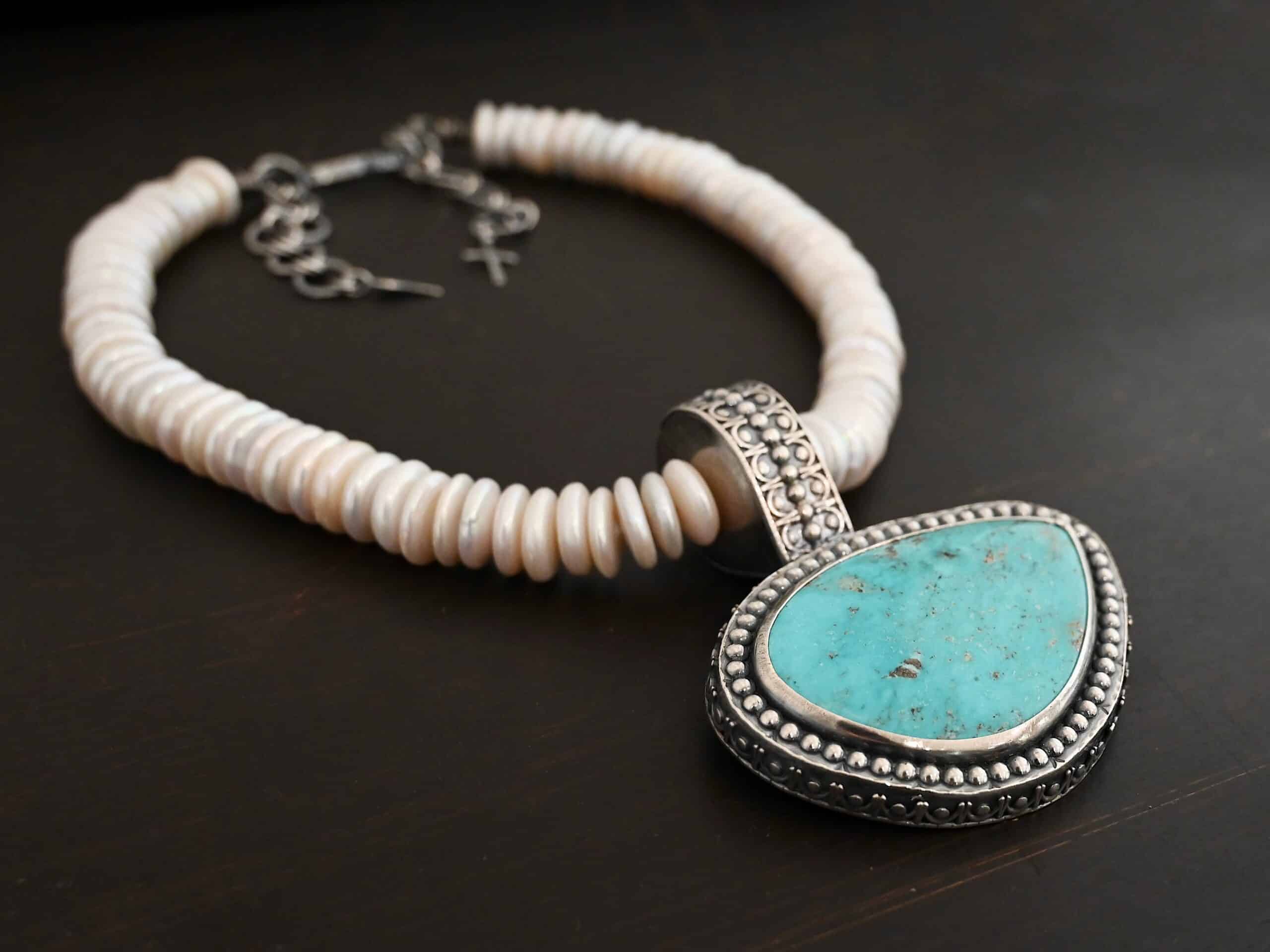 Photo of the "Cho Oyu" Necklace. It is a sterling silver, turquoise, and pearl necklace. There is a large, oval shaped turquoise set horizontally. It is set in sterling silver. The outside of the silver has silver balls set right next to each other going all the way around the turquoise. The collar of the necklace is made entirely out of strung pearl disks that are large and rough circle shapes. There is a large silver attachment on the top of the turquoise pendant to attach it to the collar. The attachment has a hole going through the center, and it is in the center of the necklace, with pearls on each side of it. Necklace is laying against a dark background.