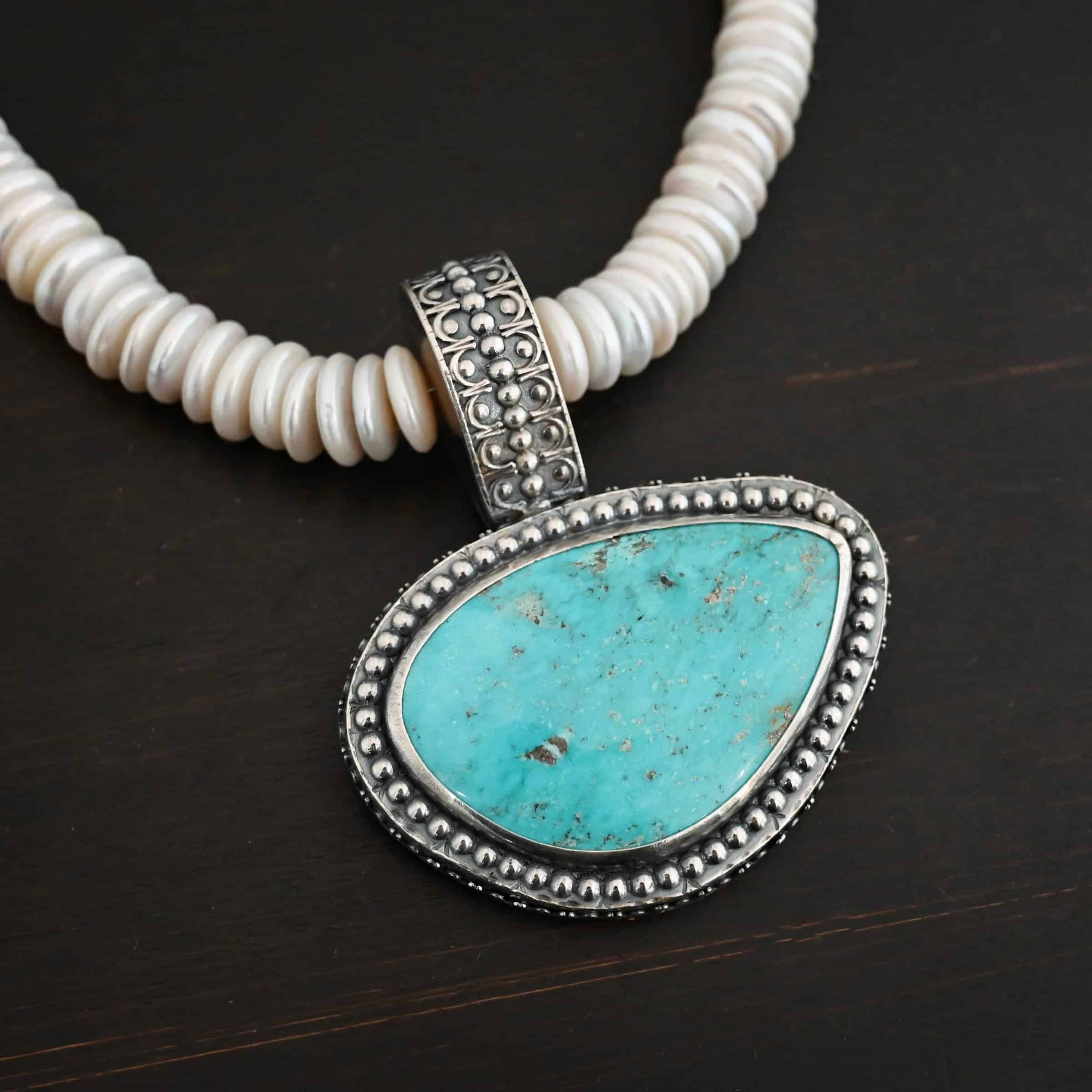 Photo of the "Cho Oyu" Necklace. It is a sterling silver, turquoise, and pearl necklace. There is a large, oval shaped turquoise set horizontally. It is set in sterling silver. The outside of the silver has silver balls set right next to each other going all the way around the turquoise. The collar of the necklace is made entirely out of strung pearl disks that are large and rough circle shapes. There is a large silver attachment on the top of the turquoise pendant to attach it to the collar. The attachment has a hole going through the center, and it is in the center of the necklace, with pearls on each side of it. Necklace is laying against a dark background.