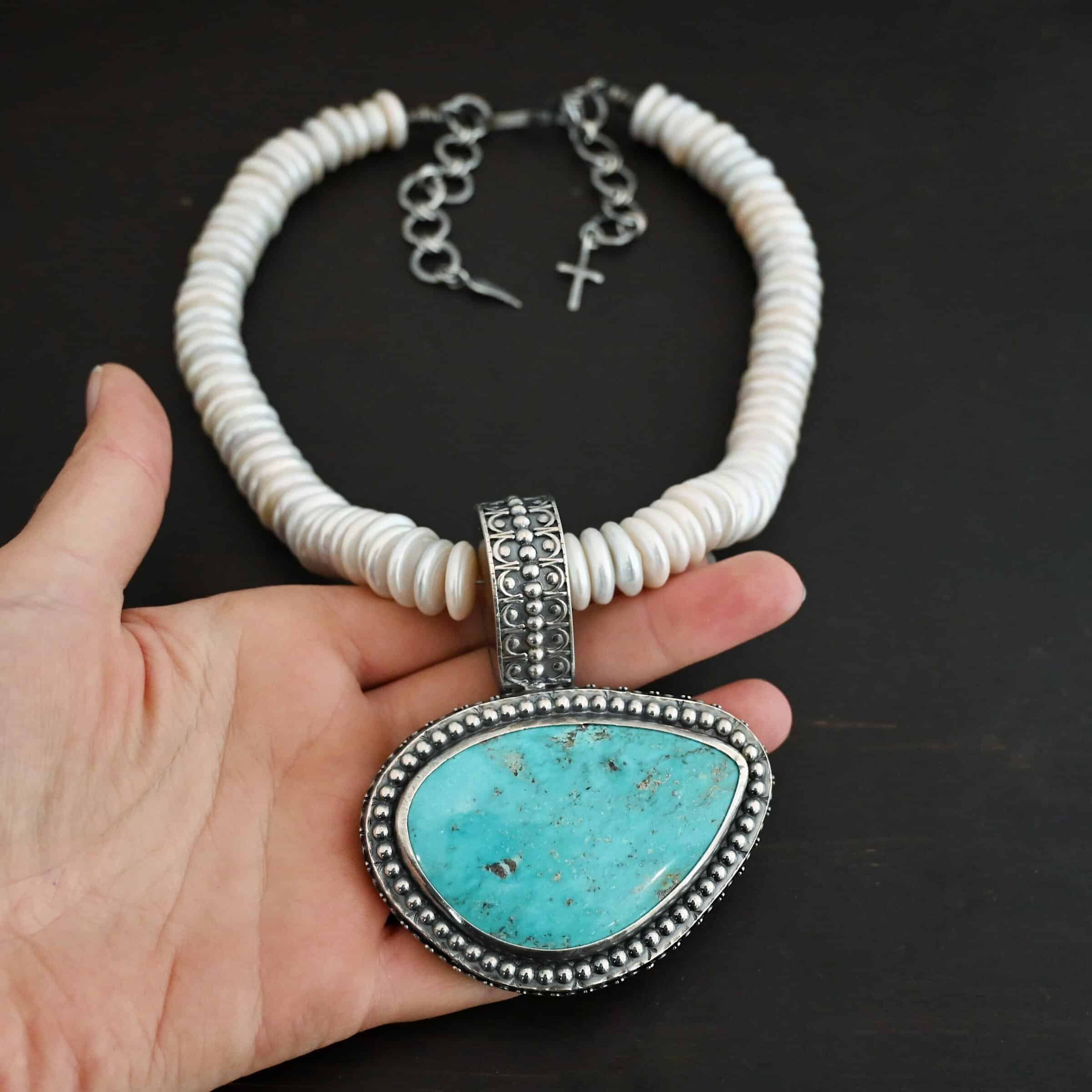 Photo of the "Cho Oyu" Necklace. It is a sterling silver, turquoise, and pearl necklace. There is a large, oval shaped turquoise set horizontally. It is set in sterling silver. The outside of the silver has silver balls set right next to each other going all the way around the turquoise. The collar of the necklace is made entirely out of strung pearl disks that are large and rough circle shapes. There is a large silver attachment on the top of the turquoise pendant to attach it to the collar. The attachment has a hole going through the center, and it is in the center of the necklace, with pearls on each side of it. Necklace is laying against the palm of a hand to demonstrate size of the pendant.
