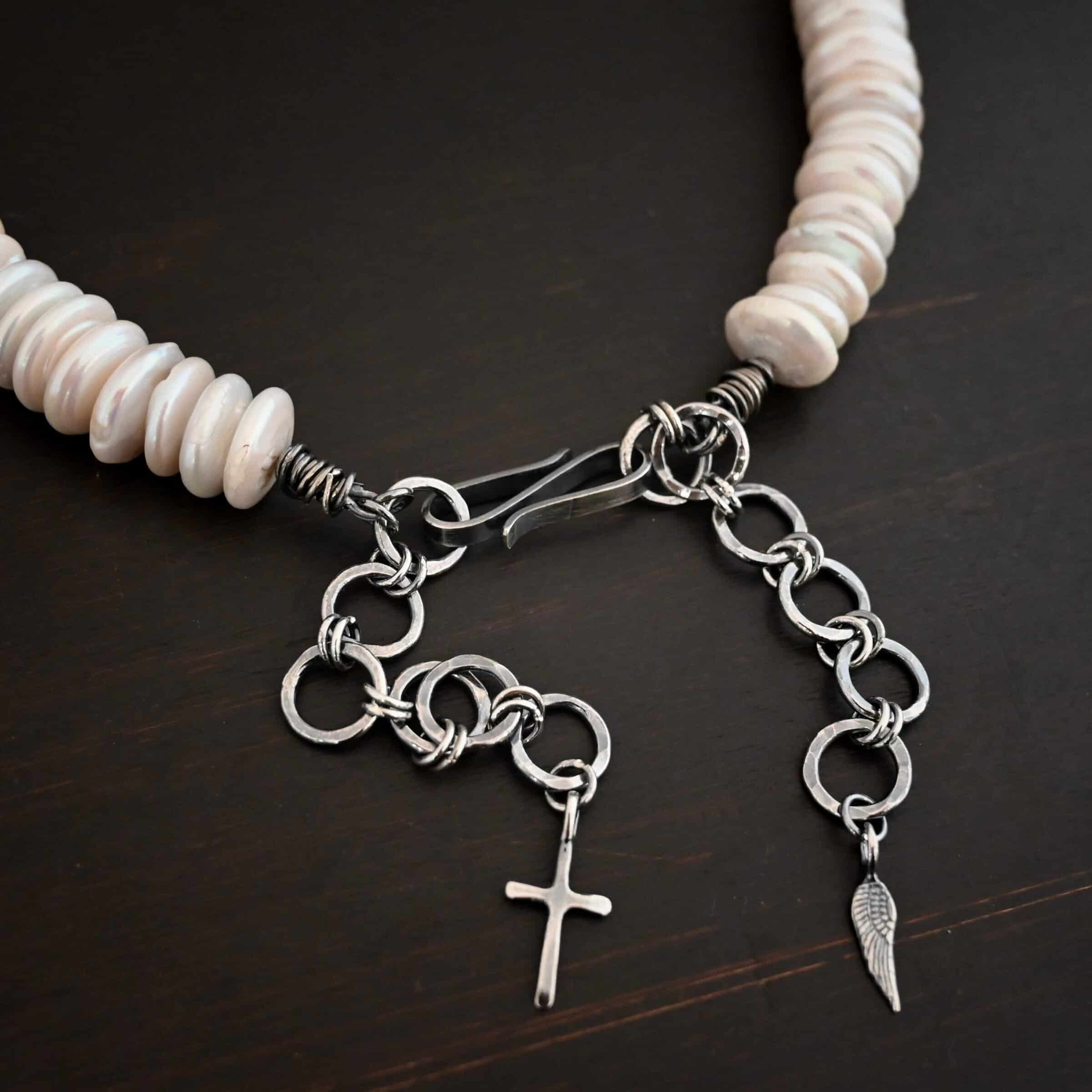 Photo of the "Cho Oyu" Necklace clasp. The collar of the necklace is made entirely out of strung pearl disks that are large and rough circle shapes. The clasp is a thick s hook clasp attached on both sides to hammered circle links that are thick for the extension of the necklace. There is a cross and angel wing charm on both ends of the extension. Clasp is laying against a dark background.