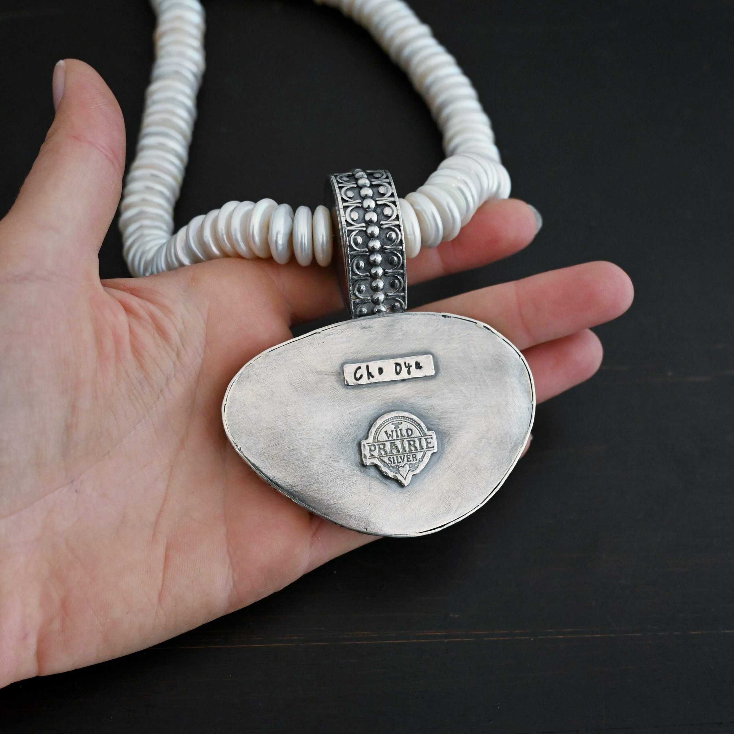Photo of the back of the "Cho Oyu" Necklace. It is a The back is entirely sterling silver, and has the words "Cho Oyu" stamped into it. The "Wild Prairie Silver" logo is also on the back, soldered to the bottom. The collar of the necklace is made entirely out of strung pearl disks that are large and rough circle shapes. There is a large silver attachment on the top of the pendant to attach it to the collar. The attachment has a hole going through the center, and it is in the center of the necklace, with pearls on each side of it. Necklace is laying against the palm of a hand to demonstrate size of the pendant.