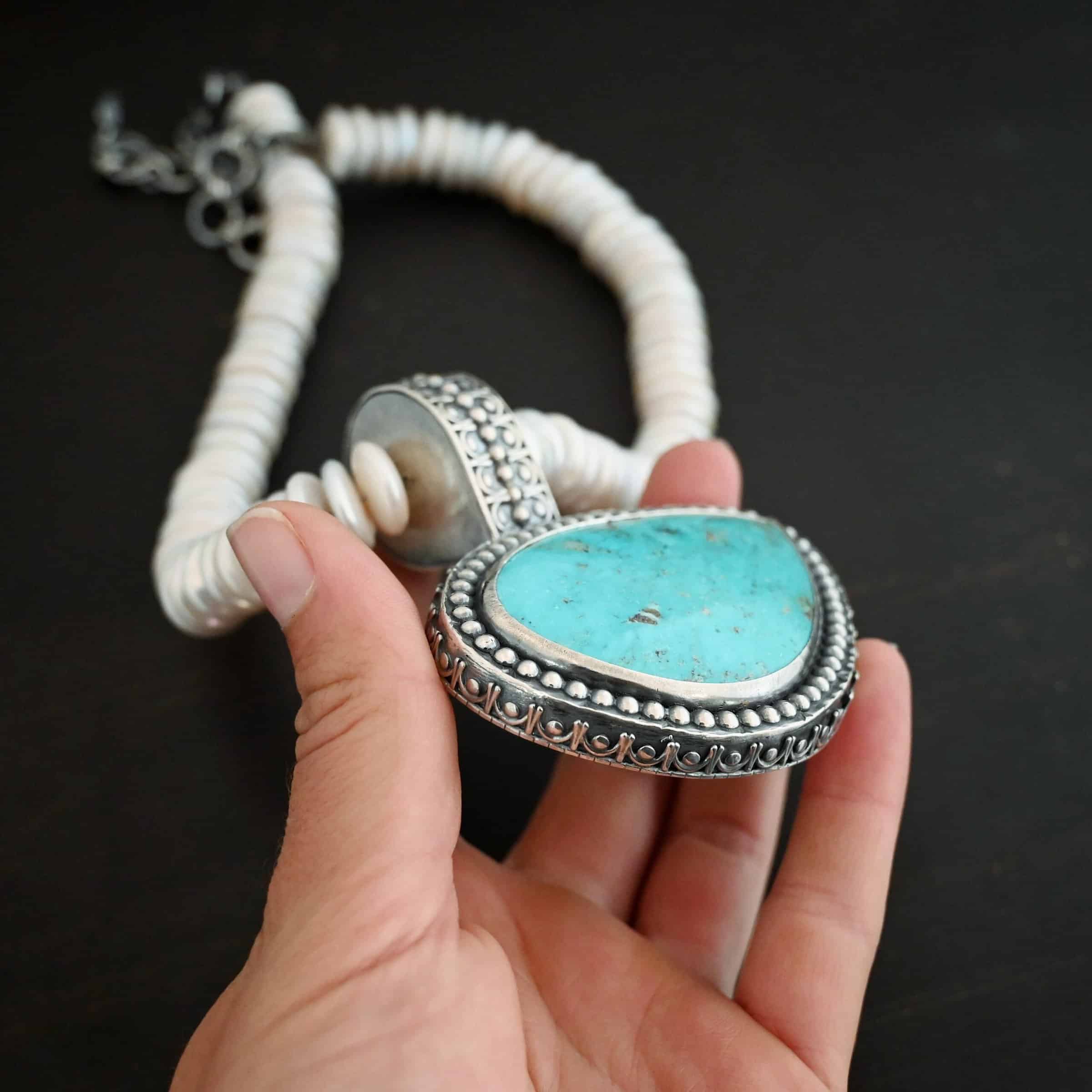 Photo of the "Cho Oyu" Necklace. It is a sterling silver, turquoise, and pearl necklace. There is a large, oval shaped turquoise set horizontally. It is set in sterling silver. The outside of the silver has silver balls set right next to each other going all the way around the turquoise. The collar of the necklace is made entirely out of strung pearl disks that are large and rough circle shapes. There is a large silver attachment on the top of the turquoise pendant to attach it to the collar. The attachment has a hole going through the center, and it is in the center of the necklace, with pearls on each side of it. Necklace is laying against the fingertips of a hand to demonstrate size of the pendant.