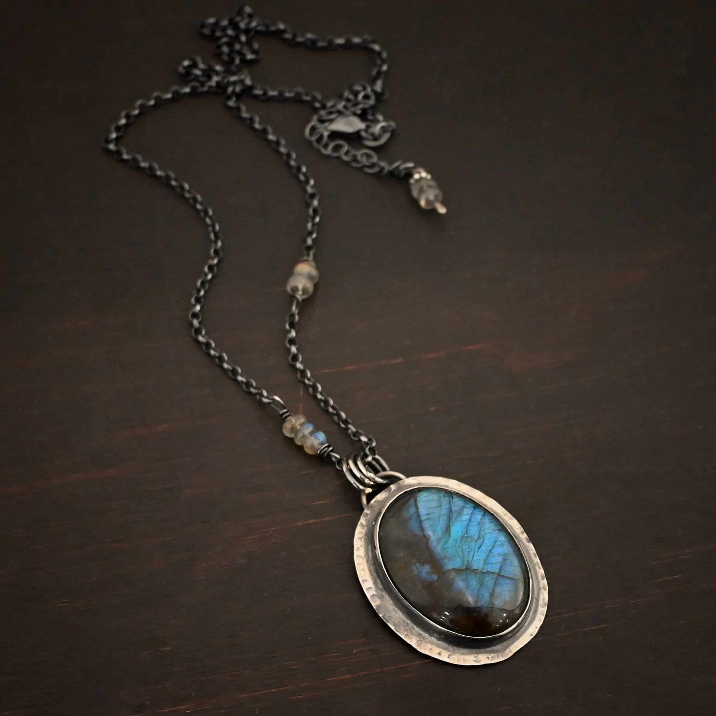 Rain Drop Necklace Close up of the "Rain Drop" Necklace. This necklace is made out of sterling silver and labradorite with a sterling silver chain that has labradorite beads on the left and right side. The beads do not hang down from the chain. Rather, it looks as if it is the chain, then the beads, and then the chain continues. The large oval labradorite is the center stone and is set in sterling silver that is hammered around the edges, and then hung onto the chain with jump rings from a loop on the back of the pendant. There are two small labradorite beads on the extension of the necklace for the chain. Necklace is laying against a dark background.