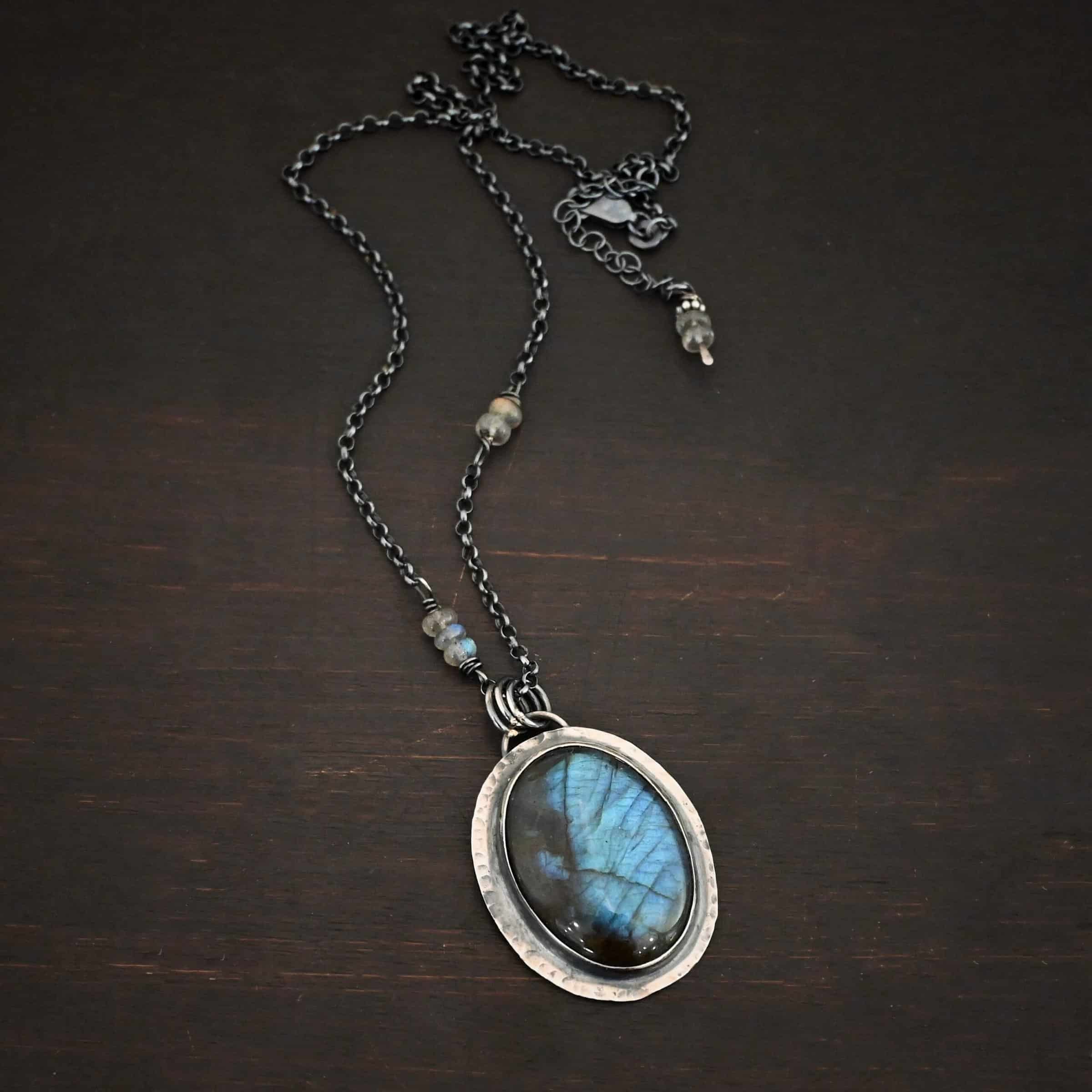 Rain Drop Necklace Close up of the "Rain Drop" Necklace. This necklace is made out of sterling silver and labradorite with a sterling silver chain that has labradorite beads on the left and right side. The beads do not hang down from the chain. Rather, it looks as if it is the chain, then the beads, and then the chain continues. The large oval labradorite is the center stone and is set in sterling silver that is hammered around the edges, and then hung onto the chain with jump rings from a loop on the back of the pendant. There are two small labradorite beads on the extension of the necklace for the chain. Necklace is laying against a dark background.