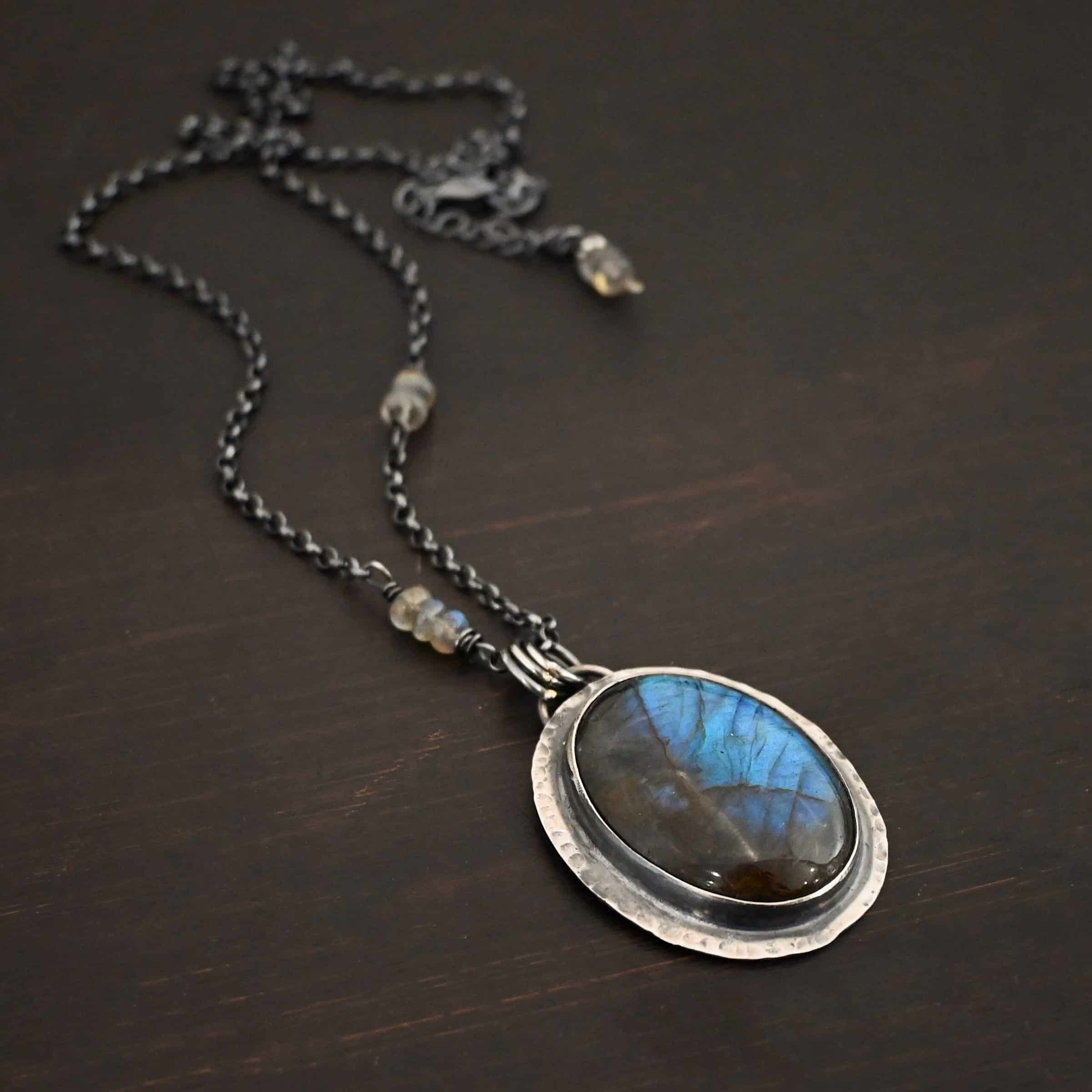 Rain Drop Necklace Close up of the "Rain Drop" Necklace. This necklace is made out of sterling silver and labradorite with a sterling silver chain that has labradorite beads on the left and right side. The beads do not hang down from the chain. Rather, it looks as if it is the chain, then the beads, and then the chain continues. The large oval labradorite is the center stone and is set in sterling silver that is hammered around the edges, and then hung onto the chain with jump rings from a loop on the back of the pendant. There are two small labradorite beads on the extension of the necklace for the chain. Necklace is laying against a dark background.