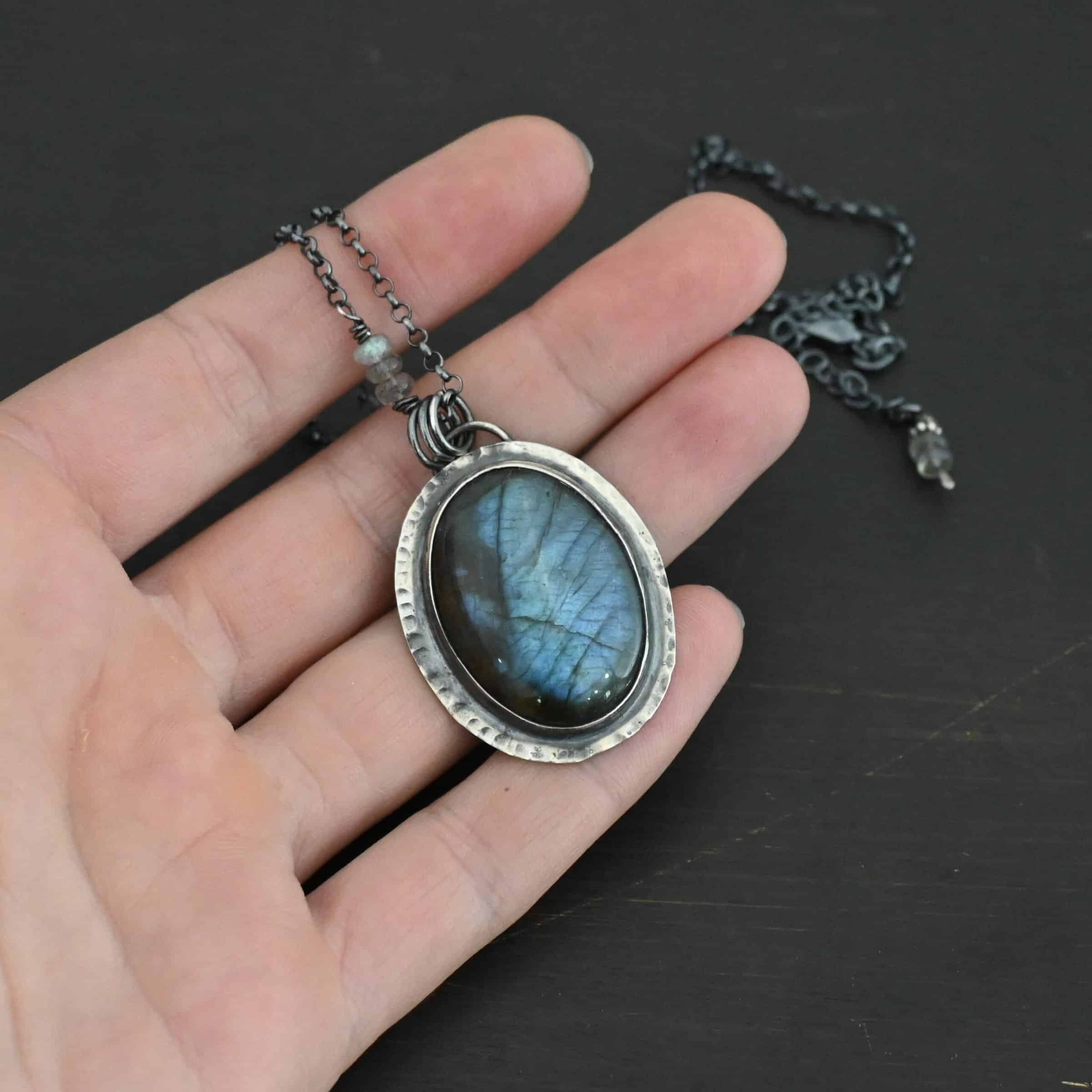 Rain Drop Necklace Close up of the "Rain Drop" Necklace. This necklace is made out of sterling silver and labradorite with a sterling silver chain that has labradorite beads on the left and right side. The beads do not hang down from the chain. Rather, it looks as if it is the chain, then the beads, and then the chain continues. The large oval labradorite is the center stone and is set in sterling silver that is hammered around the edges, and then hung onto the chain with jump rings from a loop on the back of the pendant. Necklace is laying against the fingertips of a hand to demonstrate size.