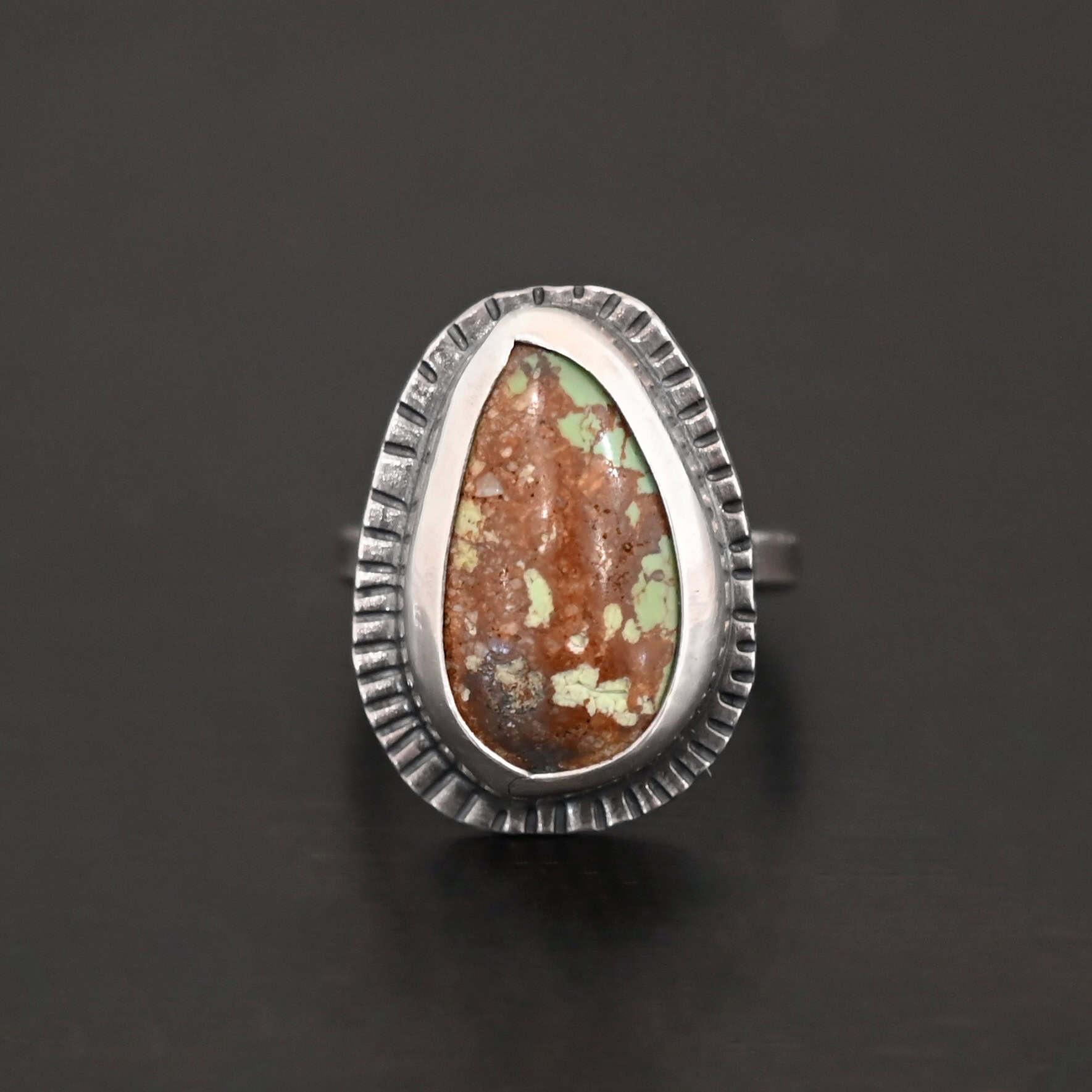 Photo of the "Anthea" Ring. Made out of sterling silver and turquoise. The turquoise is an oval shape. The outside of the ring setting has individual lines hammered all the way around the setting. The lines are darkened to stand out. Ring is sitting against a dark background.
