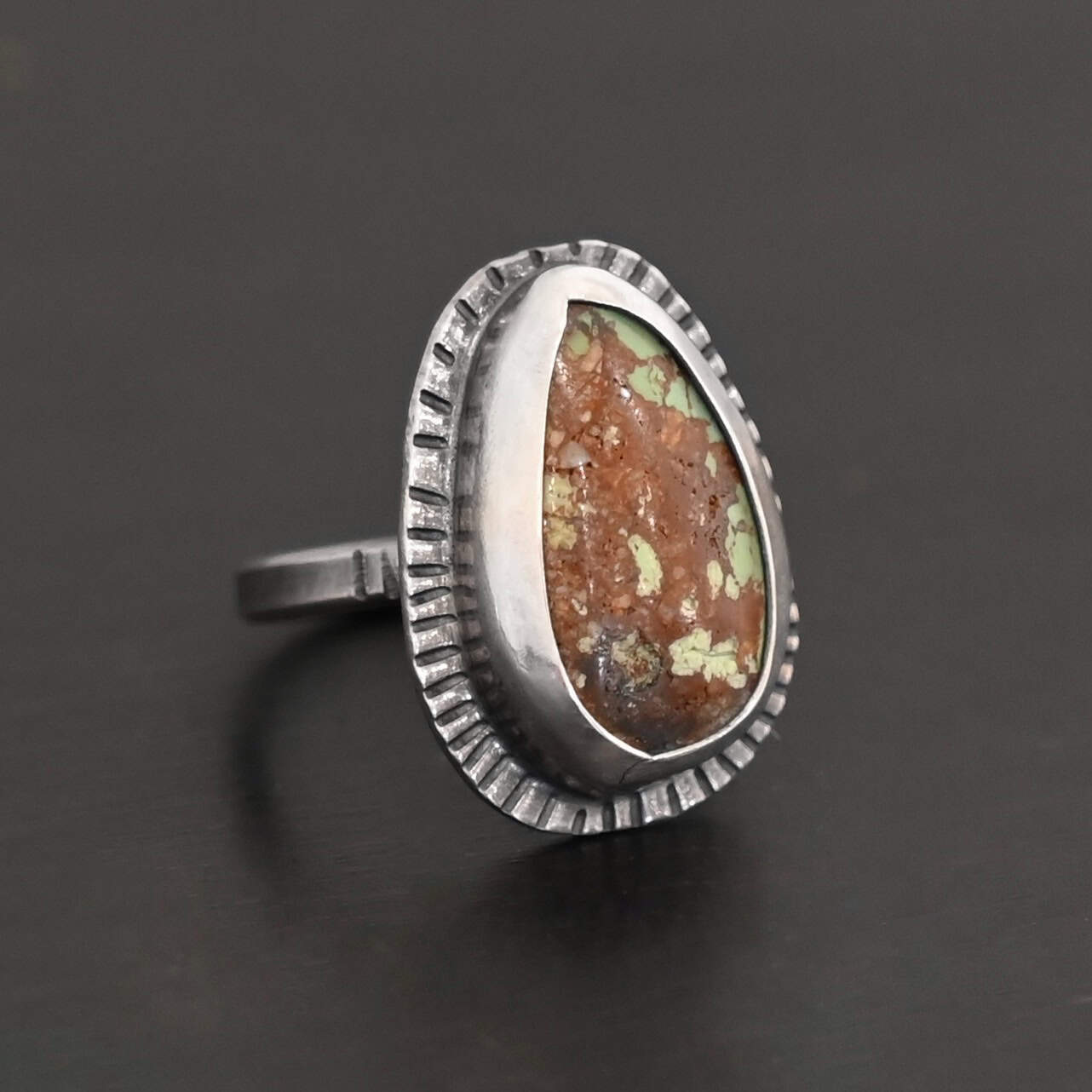 Photo of the "Anthea" Ring. Made out of sterling silver and turquoise. The turquoise is an oval shape. The outside of the ring setting has individual lines hammered all the way around the setting. The lines are darkened to stand out. Ring is sitting against a dark background.