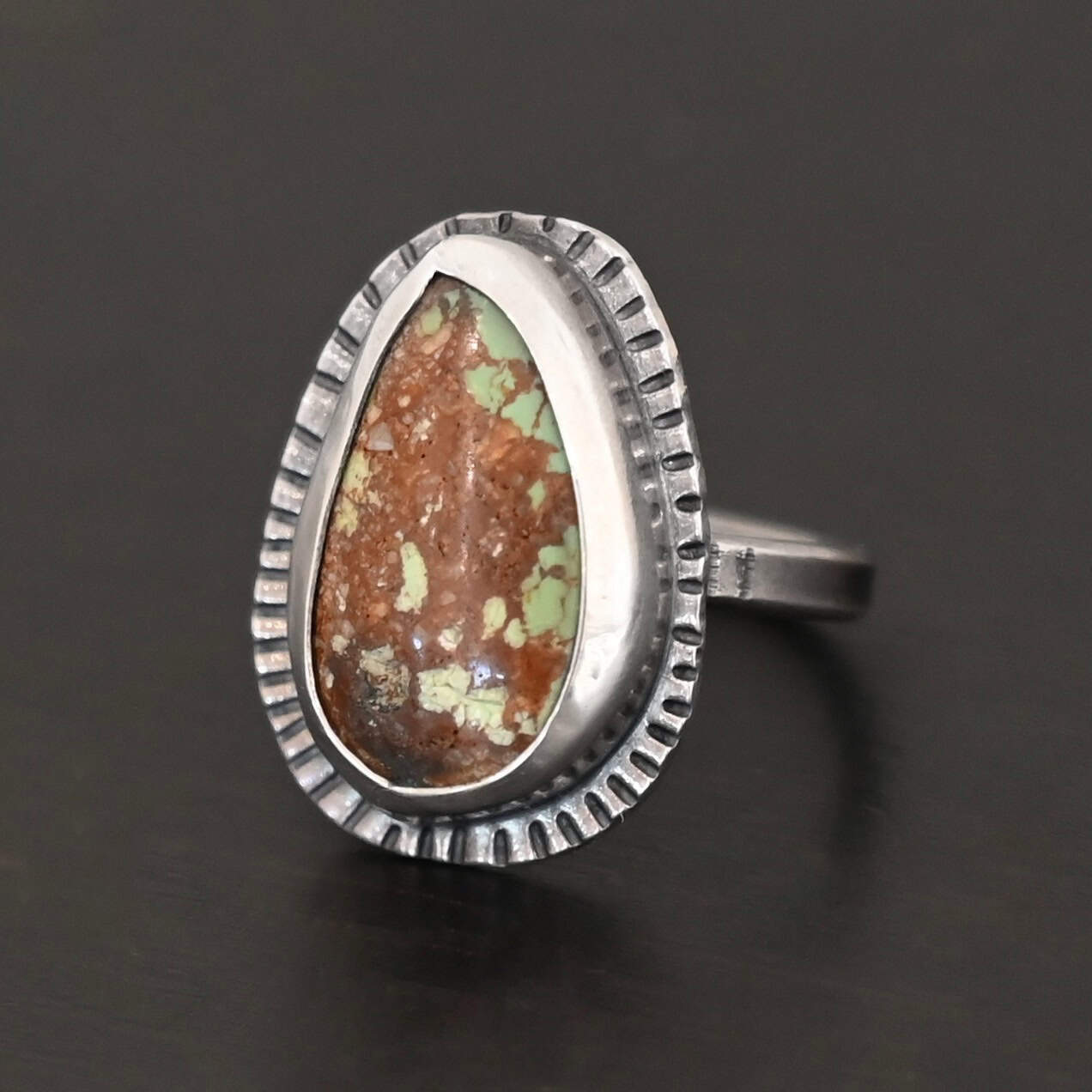 Photo of the "Anthea" Ring. Made out of sterling silver and turquoise. The turquoise is an oval shape. The outside of the ring setting has individual lines hammered all the way around the setting. The lines are darkened to stand out. Ring is sitting against a dark background.