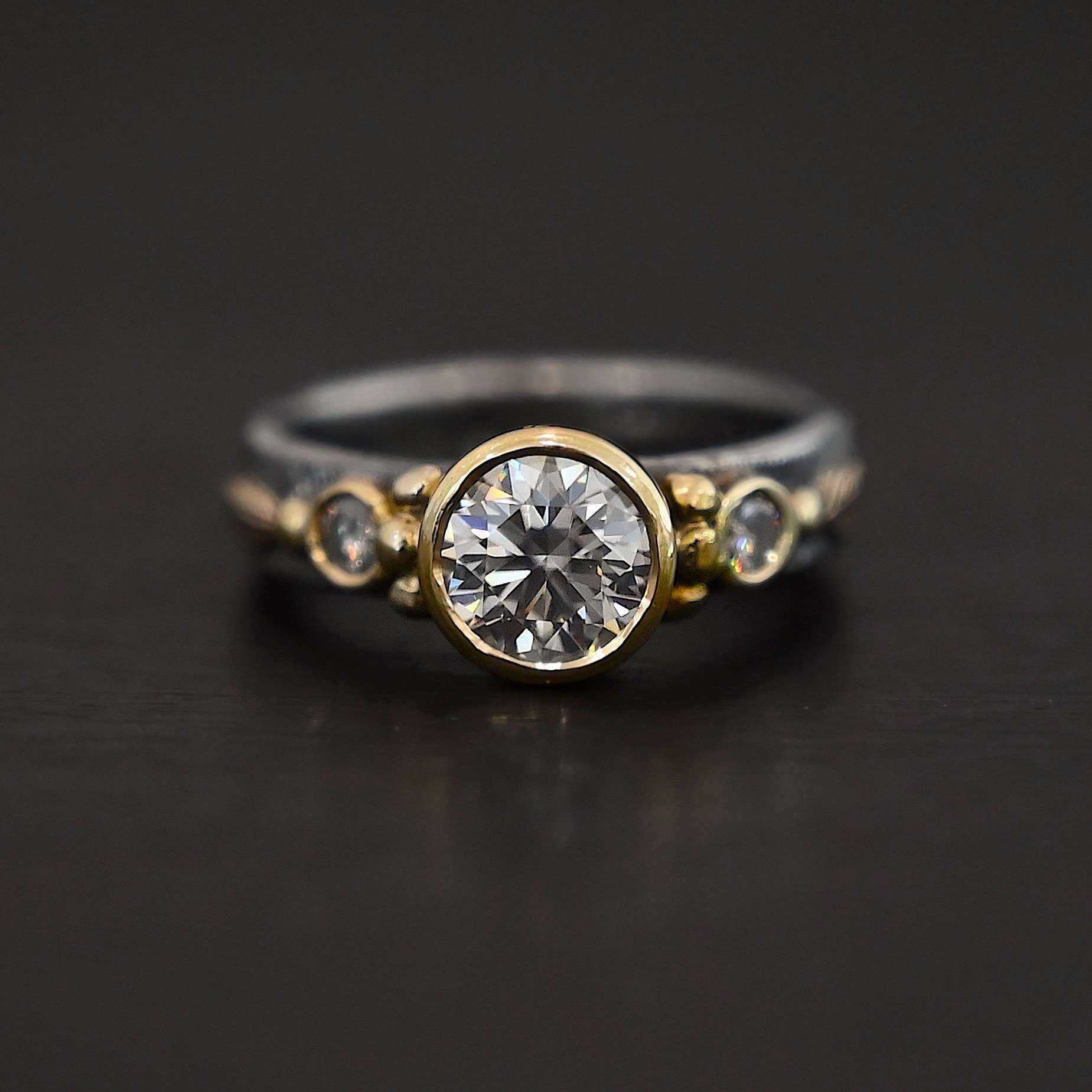 Follow Your Arrow Ring Photo of the "Follow Your Arrow" Ring. Made out of sterling silver, gold, and moissanite. There is a round moissanite in the center that is set in gold. There is a moissanite on each side of the center one. There is gold ball detail around the left and right side of the center moissanite going around the gold stone setting. Ring is sitting against a dark background.