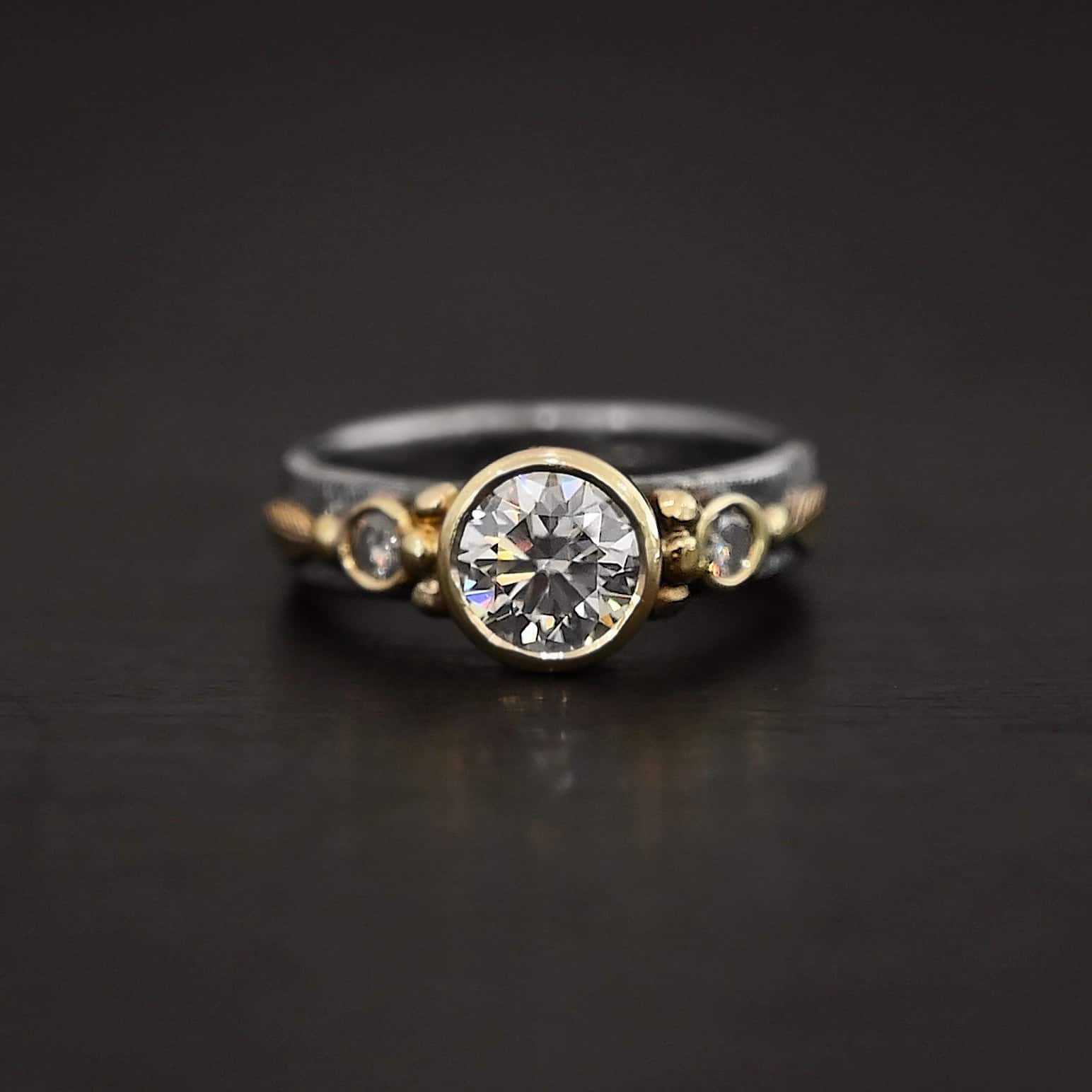 Follow Your Arrow Ring Photo of the "Follow Your Arrow" Ring. Made out of sterling silver, gold, and moissanite. There is a round moissanite in the center that is set in gold. There is a moissanite on each side of the center one. There is gold ball detail around the left and right side of the center moissanite going around the gold stone setting. Ring is sitting against a dark background.