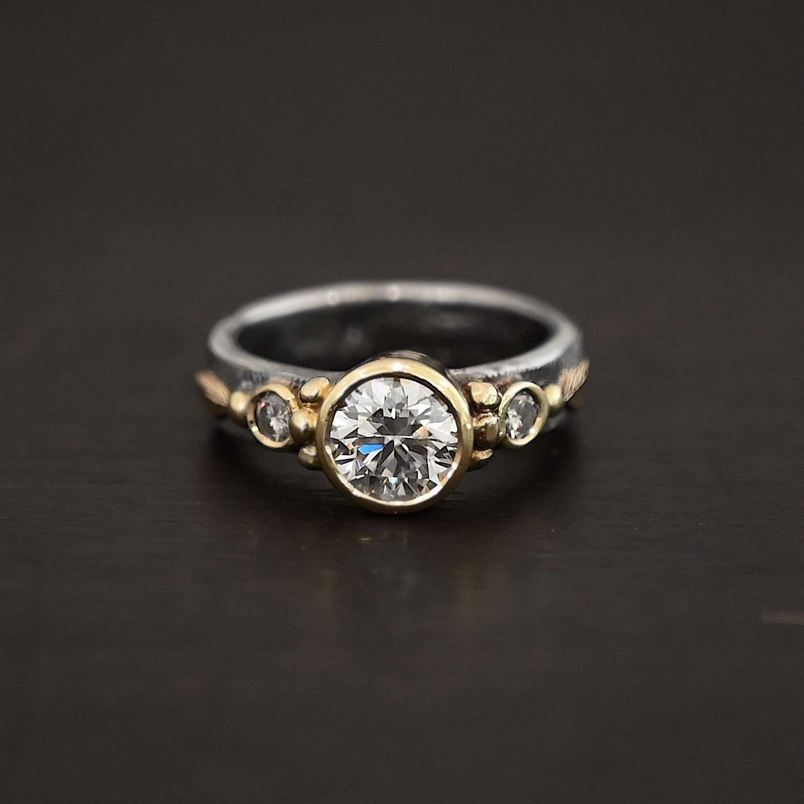 Follow Your Arrow Ring Photo of the "Follow Your Arrow" Ring. Made out of sterling silver, gold, and moissanite. There is a round moissanite in the center that is set in gold. There is a moissanite on each side of the center one. There is gold ball detail around the left and right side of the center moissanite going around the gold stone setting. Ring is sitting against a dark background.