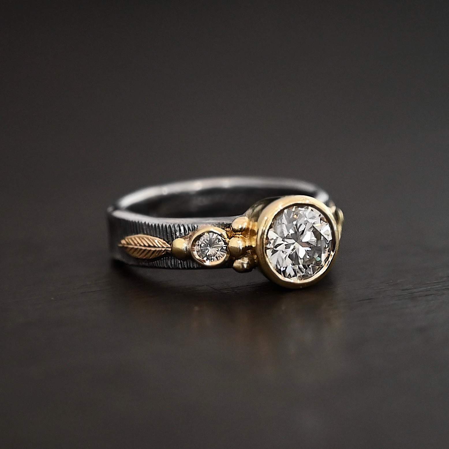 Follow Your Arrow Ring Photo of the "Follow Your Arrow" Ring. Made out of sterling silver, gold, and moissanite. There is a round moissanite in the center that is set in gold. There is a moissanite on each side of the center one. There is gold ball detail around the left and right side of the center moissanite going around the gold stone setting. Ring is sitting against a dark background.