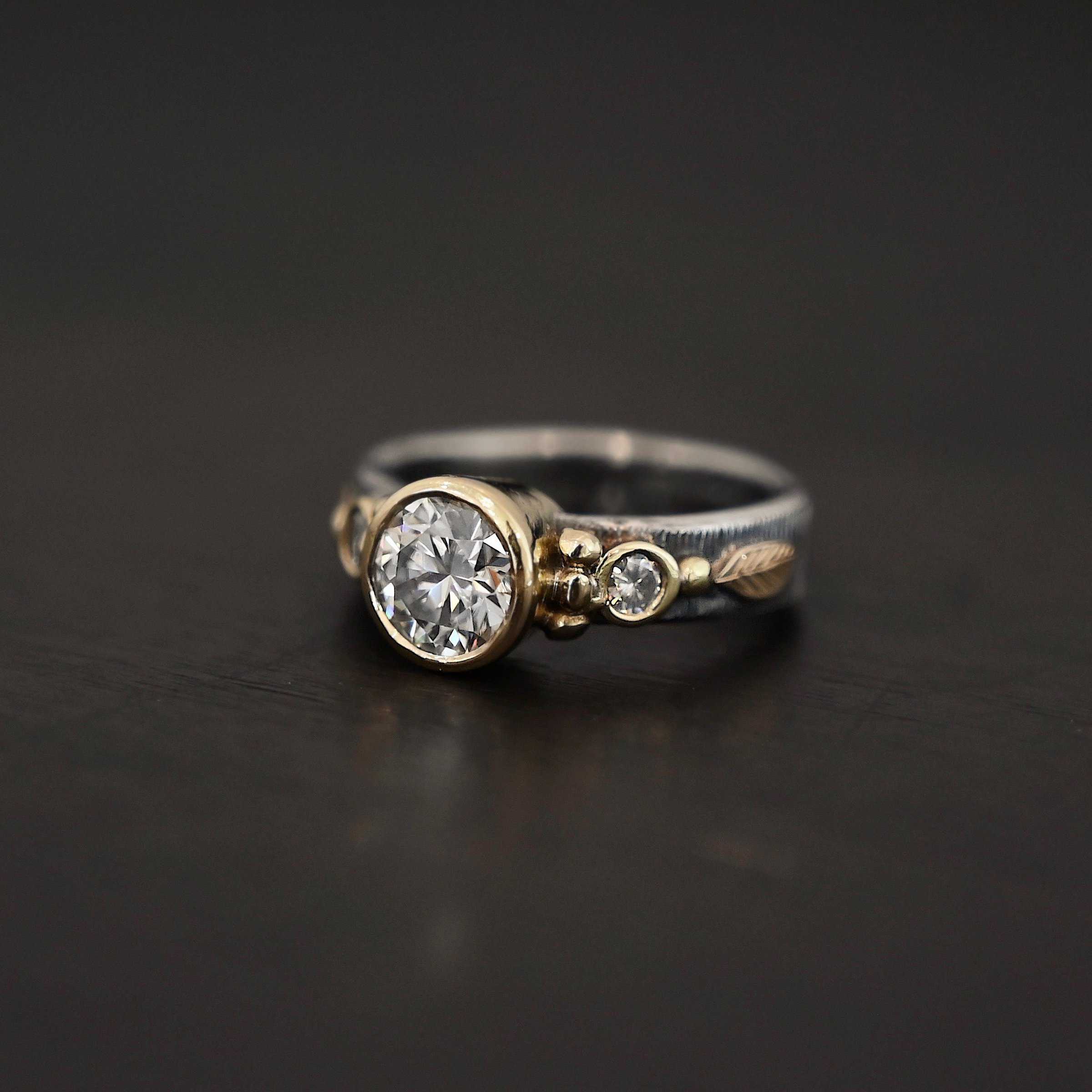 Follow Your Arrow Ring Photo of the "Follow Your Arrow" Ring. Made out of sterling silver, gold, and moissanite. There is a round moissanite in the center that is set in gold. There is a moissanite on each side of the center one. There is gold ball detail around the left and right side of the center moissanite going around the gold stone setting. Ring is sitting against a dark background.