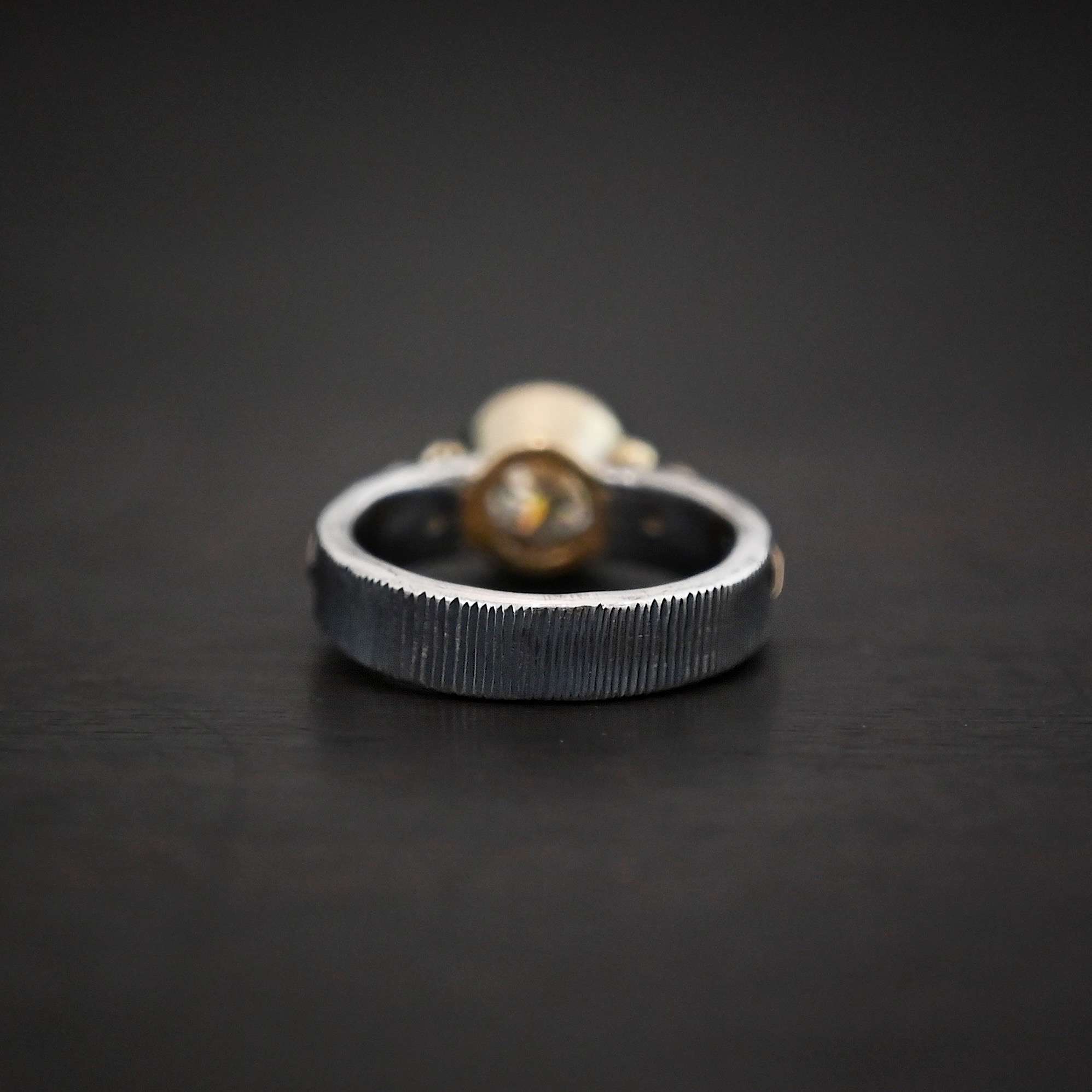 Follow Your Arrow Ring Photo of the back of the "Follow Your Arrow" Ring. Made out of sterling silver and gold. The ring band is circular. The back of the ring has straight lines carved into it. The lines are very close together. Ring is sitting against a dark background.