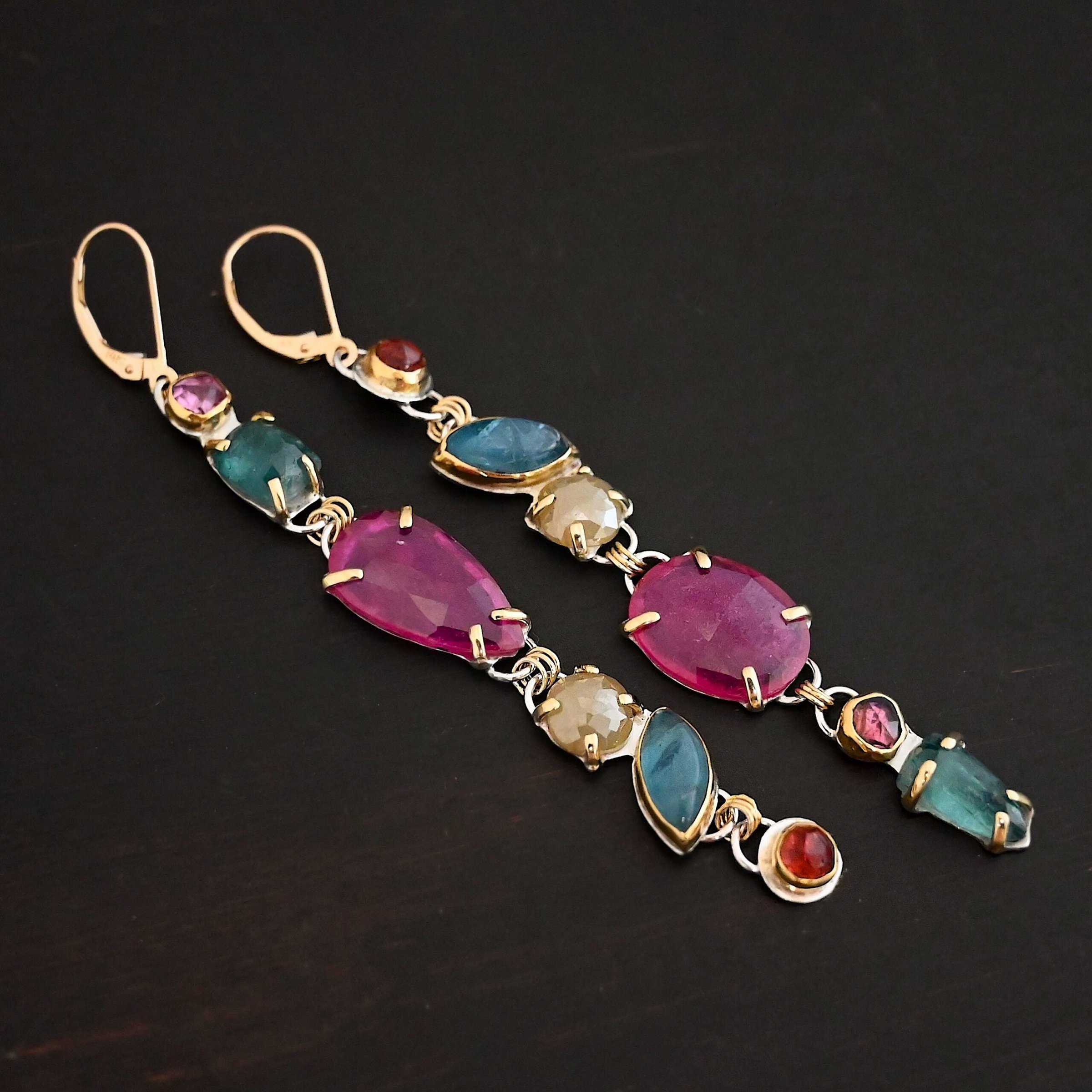 Follow the Path Earrings Photo of the "Follow Your Path" Earrings. Made out of sterling silver & 18K gold, tourmalines, apatites, rough diamonds, & rubies. They are approximately 3.5″ long. The rubies are in the center, and the rest of the stones are above or beneath them. The rubies are held in with gold prongs, as well as some of the other stones. Each stone setting has a loop on the back of it with gold jump rings that connect the stones to one another. Earrings have gold leverback ear wires. Earrings are laying against a dark background.