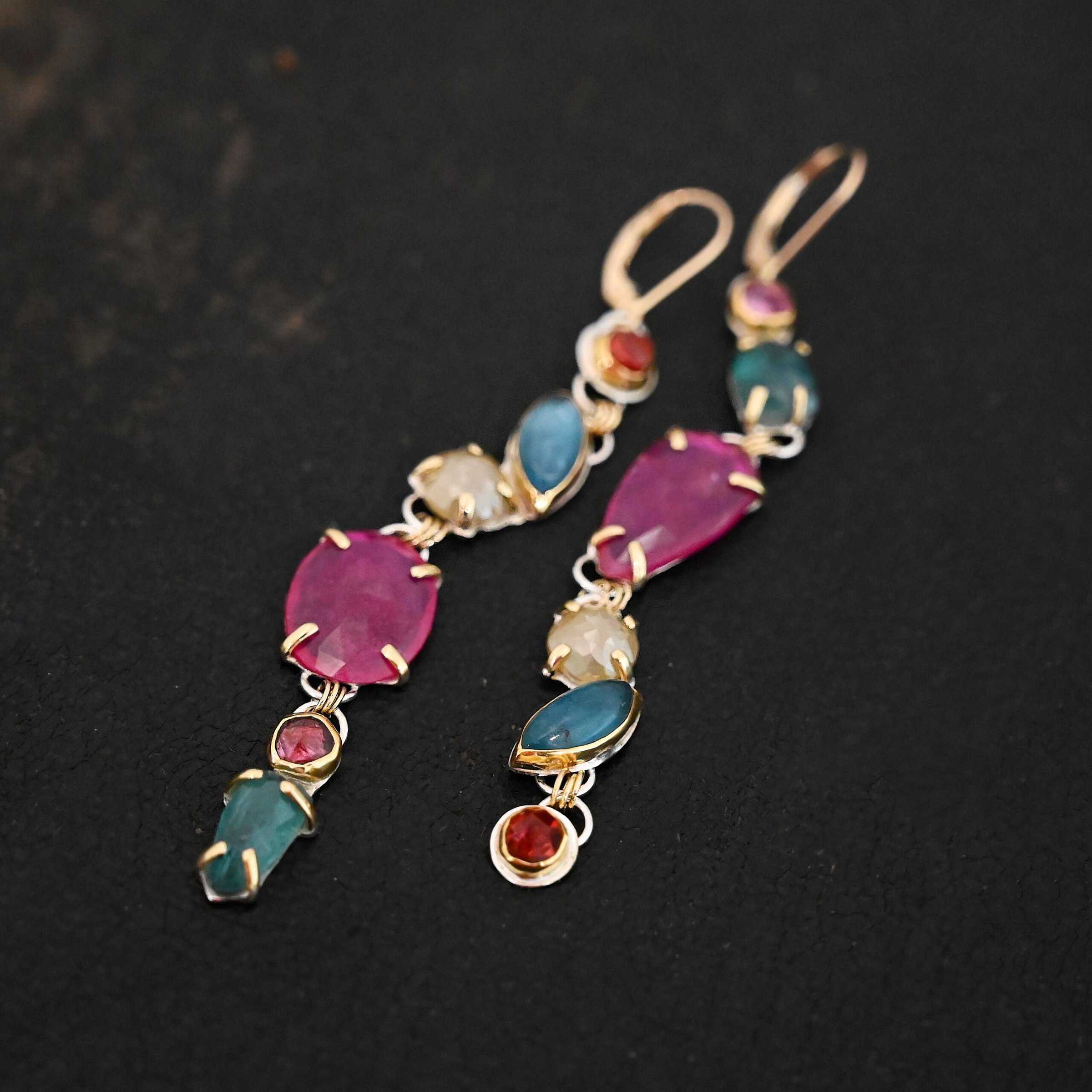 Follow the Path Earrings Photo of the "Follow Your Path" Earrings. Made out of sterling silver & 18K gold, tourmalines, apatites, rough diamonds, & rubies. They are approximately 3.5″ long. The rubies are in the center, and the rest of the stones are above or beneath them. The rubies are held in with gold prongs, as well as some of the other stones. Each stone setting has a loop on the back of it with gold jump rings that connect the stones to one another. Earrings have gold leverback ear wires. Earrings are laying against a dark background.