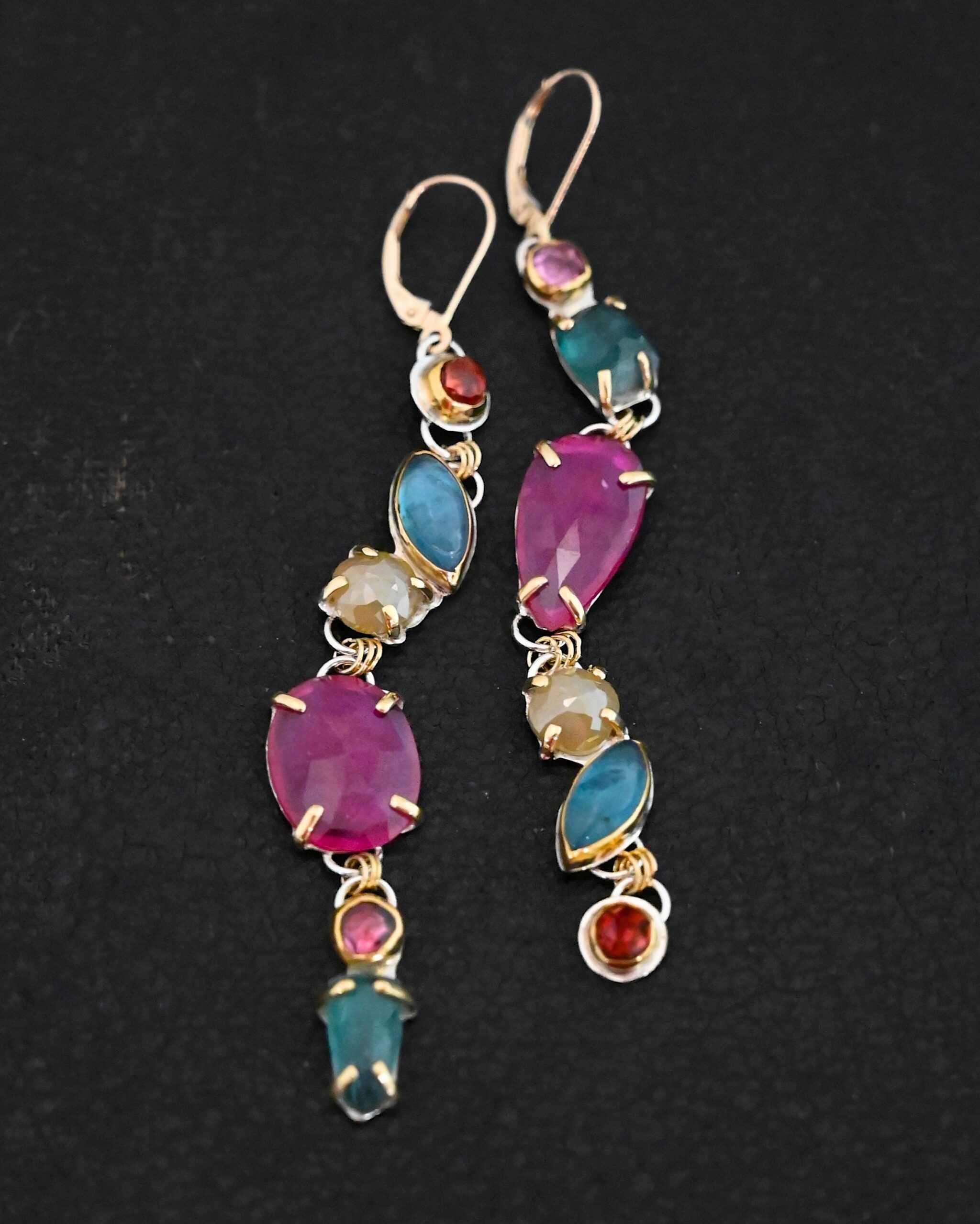 Follow the Path Earrings Photo of the "Follow Your Path" Earrings. Made out of sterling silver & 18K gold, tourmalines, apatites, rough diamonds, & rubies. They are approximately 3.5″ long. The rubies are in the center, and the rest of the stones are above or beneath them. The rubies are held in with gold prongs, as well as some of the other stones. Each stone setting has a loop on the back of it with gold jump rings that connect the stones to one another. Earrings have gold leverback ear wires. Earrings are laying against a dark background.