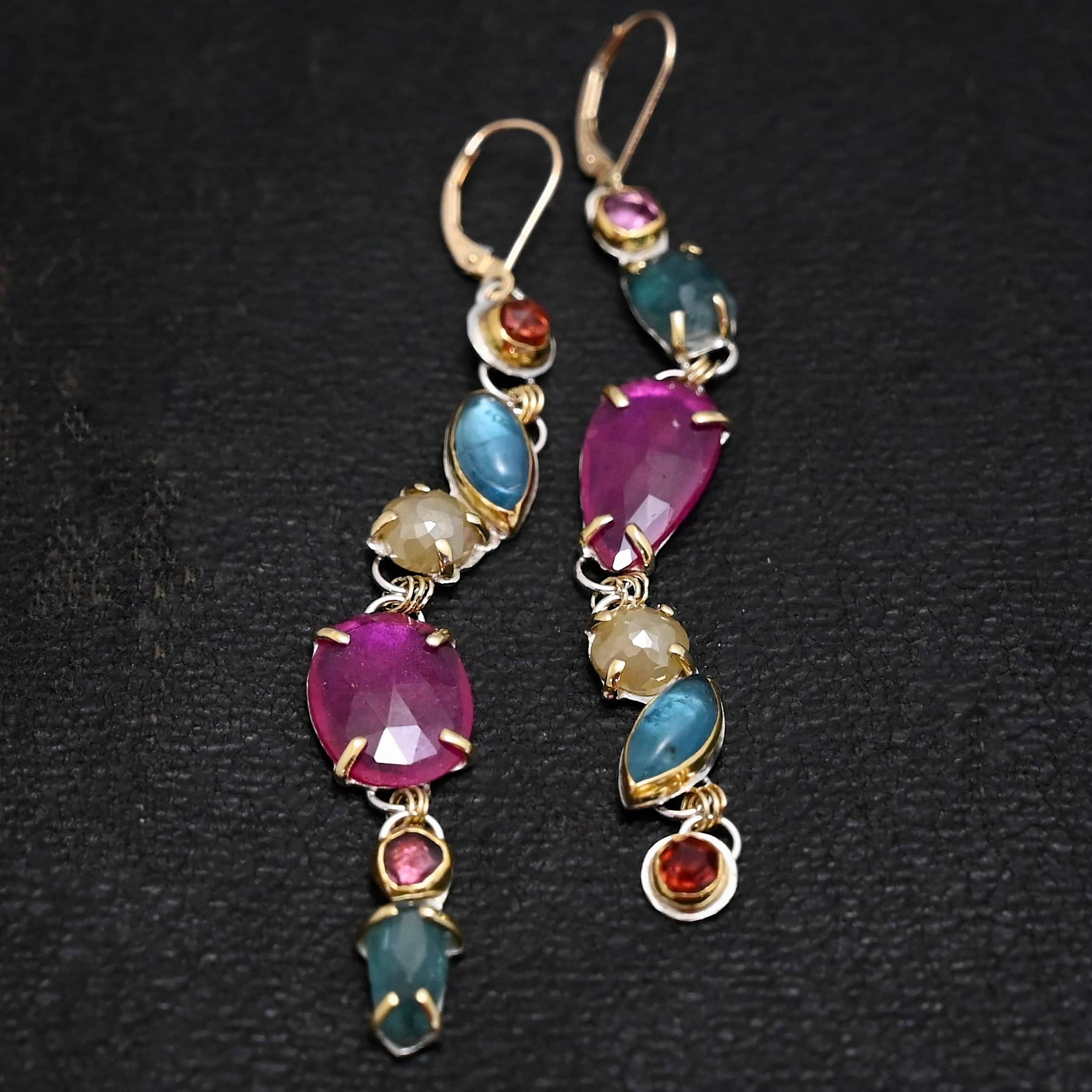 Follow the Path Earrings Photo of the "Follow Your Path" Earrings. Made out of sterling silver & 18K gold, tourmalines, apatites, rough diamonds, & rubies. They are approximately 3.5″ long. The rubies are in the center, and the rest of the stones are above or beneath them. The rubies are held in with gold prongs, as well as some of the other stones. Each stone setting has a loop on the back of it with gold jump rings that connect the stones to one another. Earrings have gold leverback ear wires. Earrings are laying against a dark background.