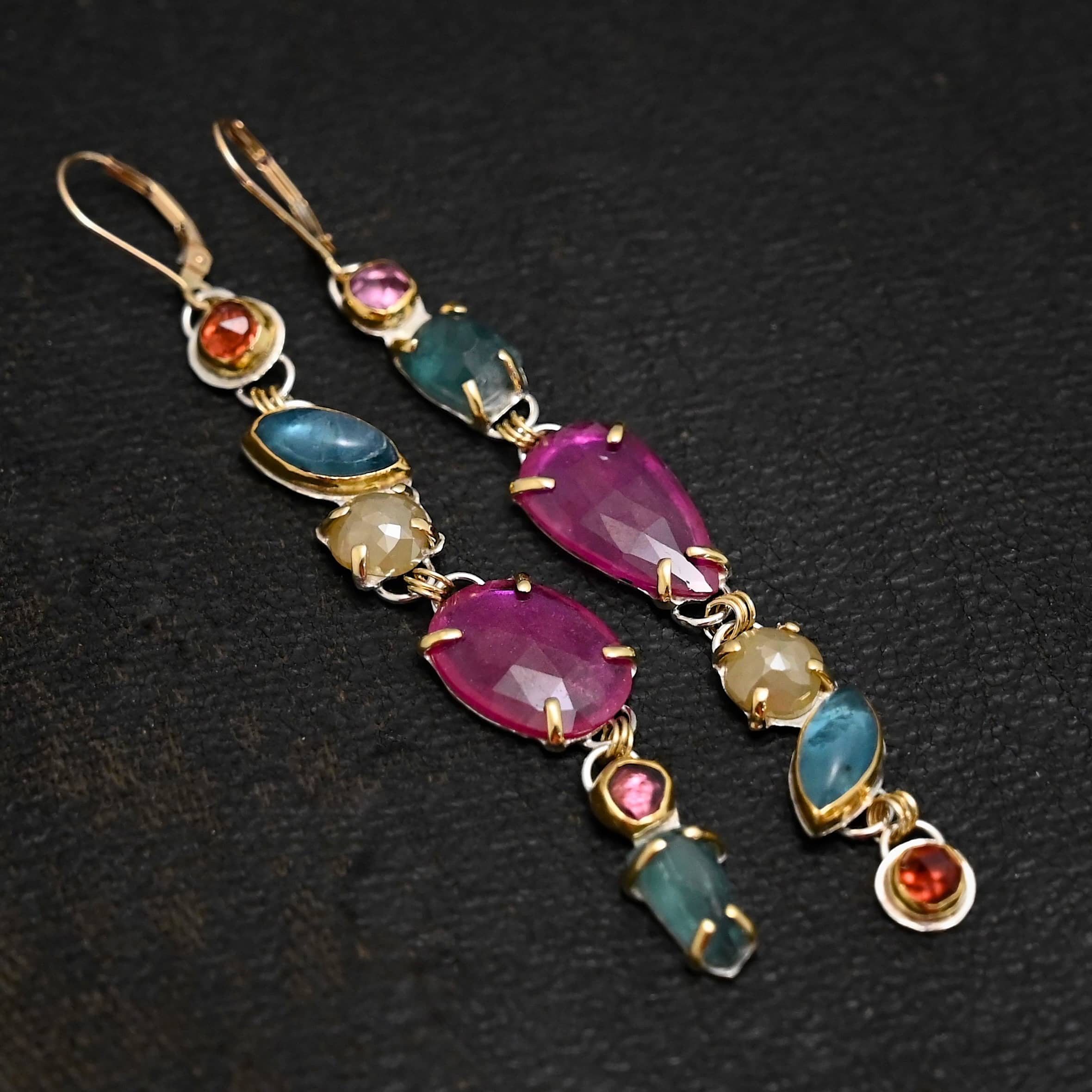 Follow the Path Earrings Photo of the "Follow Your Path" Earrings. Made out of sterling silver & 18K gold, tourmalines, apatites, rough diamonds, & rubies. They are approximately 3.5″ long. The rubies are in the center, and the rest of the stones are above or beneath them. The rubies are held in with gold prongs, as well as some of the other stones. Each stone setting has a loop on the back of it with gold jump rings that connect the stones to one another. Earrings have gold leverback ear wires. Earrings are laying against a dark background.