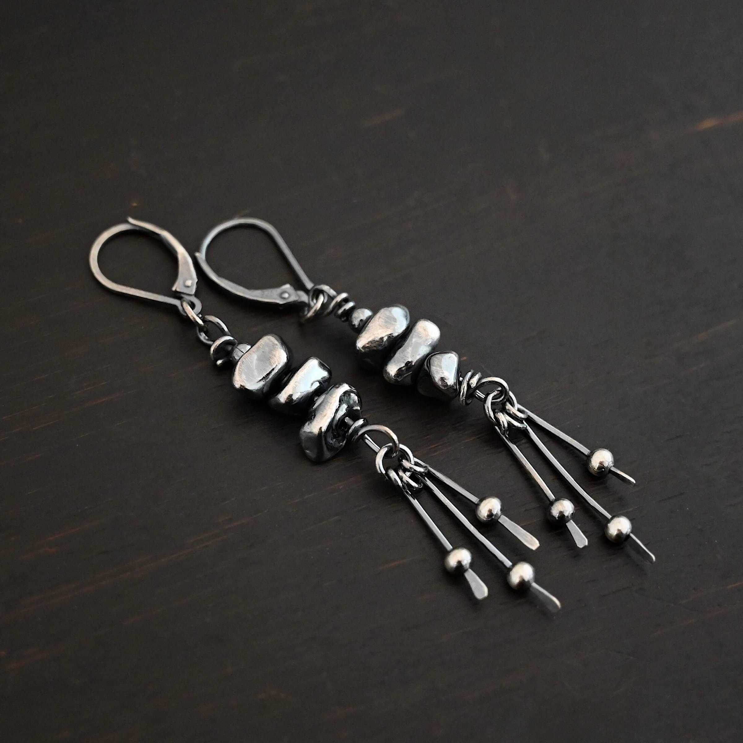 Photo of the "Pebble Beach" Earrings. Made out of sterling silver. The earrings have leverback ear wires. Each earring has three silver pebbles on top of one another beneath the ear wires. Hanging from the bottom of the pebbles are three short silver sticks with the ends hammered into paddles. Above the paddles are silver balls. There is one silver ball on each paddle. Earrings are laying against a dark background.