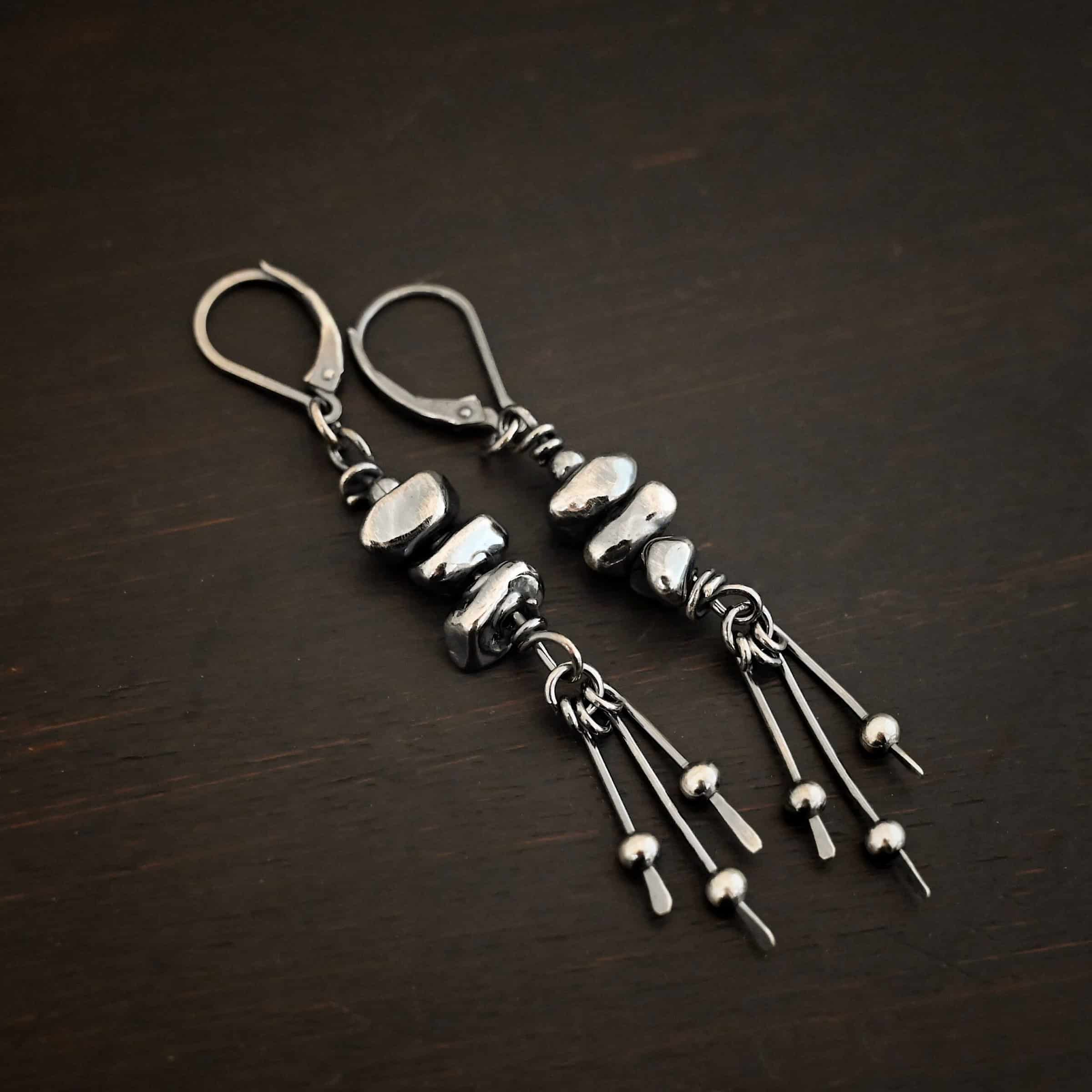 Photo of the "Pebble Beach" Earrings. Made out of sterling silver. The earrings have leverback ear wires. Each earring has three silver pebbles on top of one another beneath the ear wires. Hanging from the bottom of the pebbles are three short silver sticks with the ends hammered into paddles. Above the paddles are silver balls. There is one silver ball on each paddle. Earrings are laying against a dark background.
