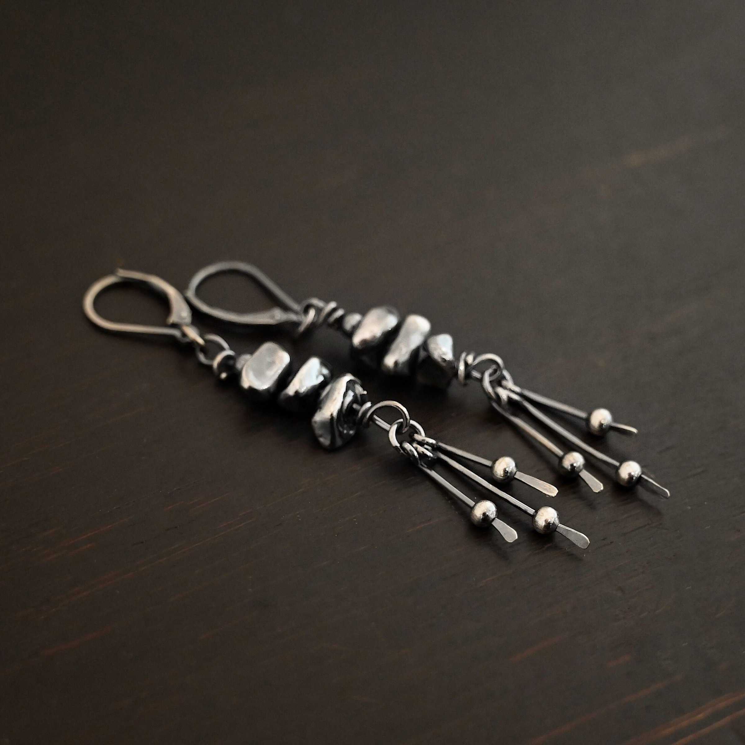 Photo of the "Pebble Beach" Earrings. Made out of sterling silver. The earrings have leverback ear wires. Each earring has three silver pebbles on top of one another beneath the ear wires. Hanging from the bottom of the pebbles are three short silver sticks with the ends hammered into paddles. Above the paddles are silver balls. There is one silver ball on each paddle. Earrings are laying against a dark background.