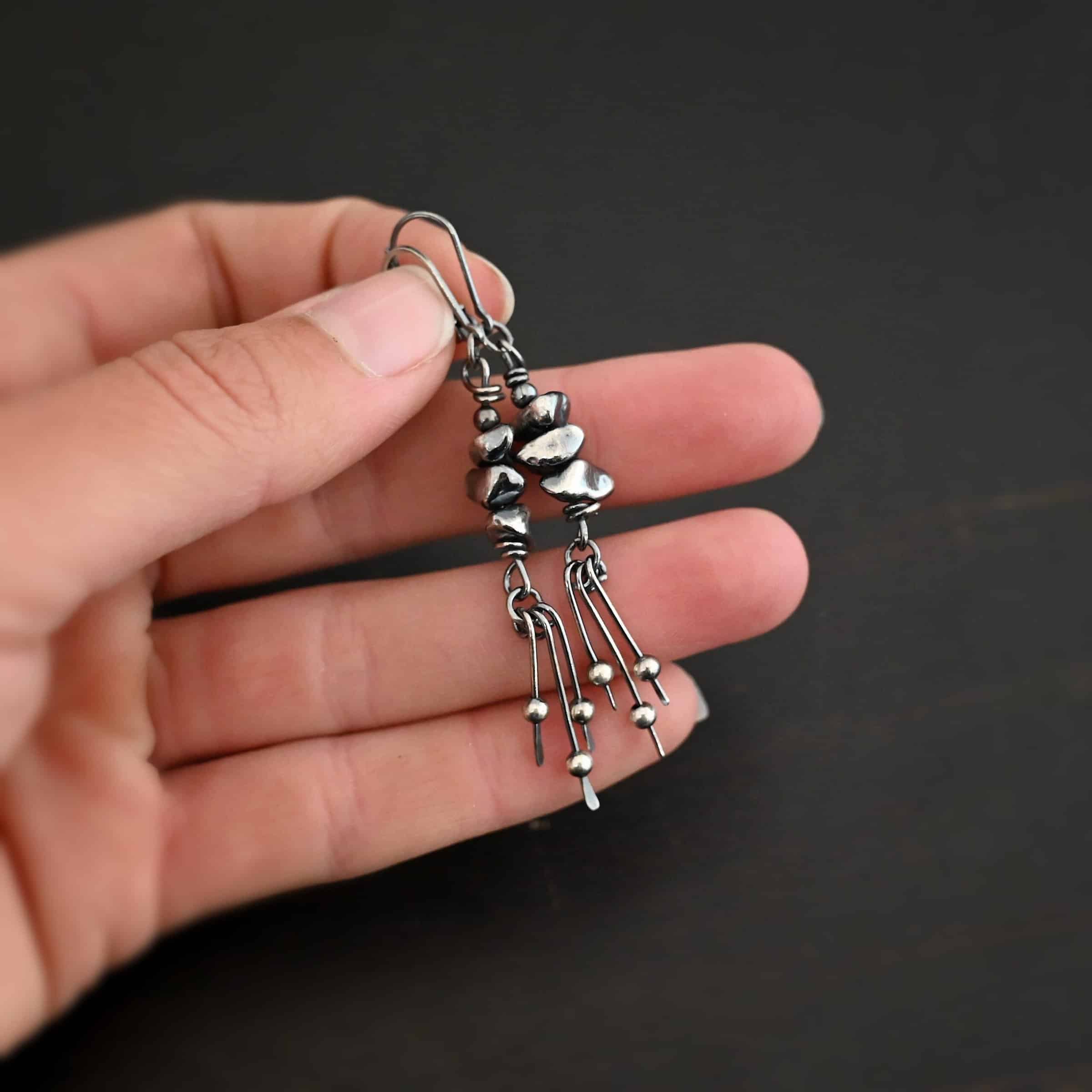 Photo of the "Pebble Beach" Earrings. Made out of sterling silver. The earrings have leverback ear wires. Each earring has three silver pebbles on top of one another beneath the ear wires. Hanging from the bottom of the pebbles are three short silver sticks with the ends hammered into paddles. Above the paddles are silver balls. There is one silver ball on each paddle. Earrings are held in the fingertips of a hand to demonstrate size.