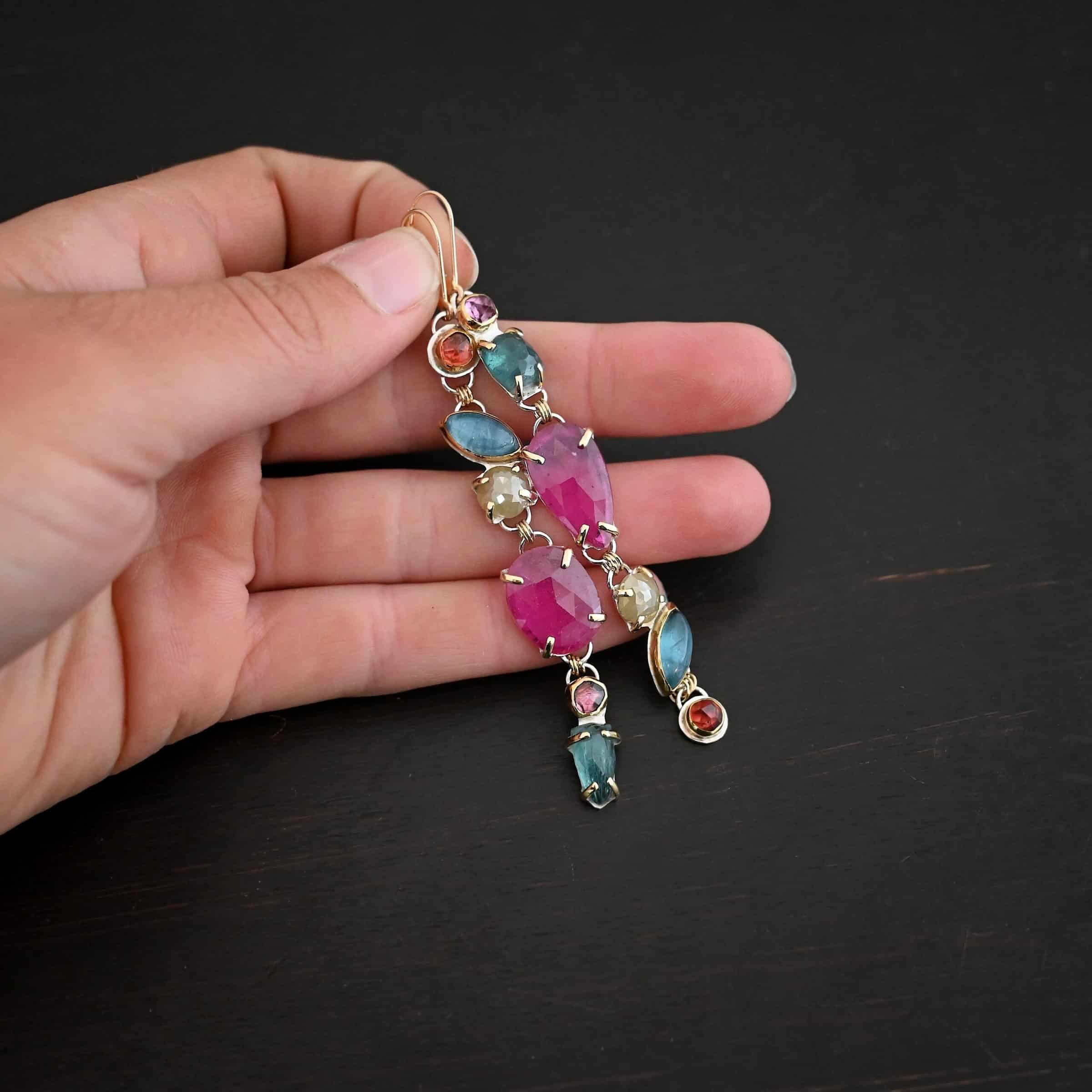 Follow the Path Earrings Photo of the "Follow Your Path" Earrings. Made out of sterling silver & 18K gold, tourmalines, apatites, rough diamonds, & rubies. They are approximately 3.5″ long. The rubies are in the center, and the rest of the stones are above or beneath them. The rubies are held in with gold prongs, as well as some of the other stones. Each stone setting has a loop on the back of it with gold jump rings that connect the stones to one another. Earrings have gold leverback ear wires. Earrings are laying against the fingertips of a hand to demonstrate size.