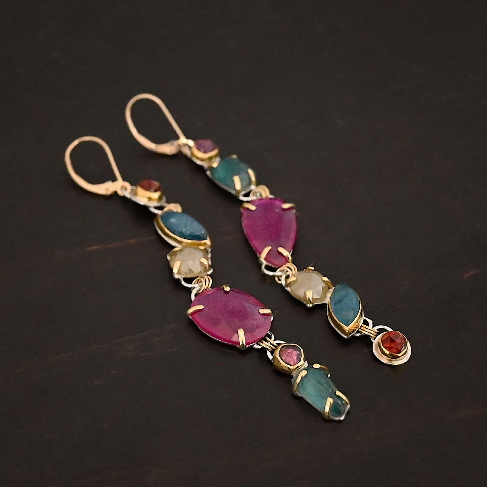Follow the Path Earrings Photo of the "Follow Your Path" Earrings. Made out of sterling silver & 18K gold, tourmalines, apatites, rough diamonds, & rubies. They are approximately 3.5″ long. The rubies are in the center, and the rest of the stones are above or beneath them. The rubies are held in with gold prongs, as well as some of the other stones. Each stone setting has a loop on the back of it with gold jump rings that connect the stones to one another. Earrings have gold leverback ear wires. Earrings are laying against a dark background.