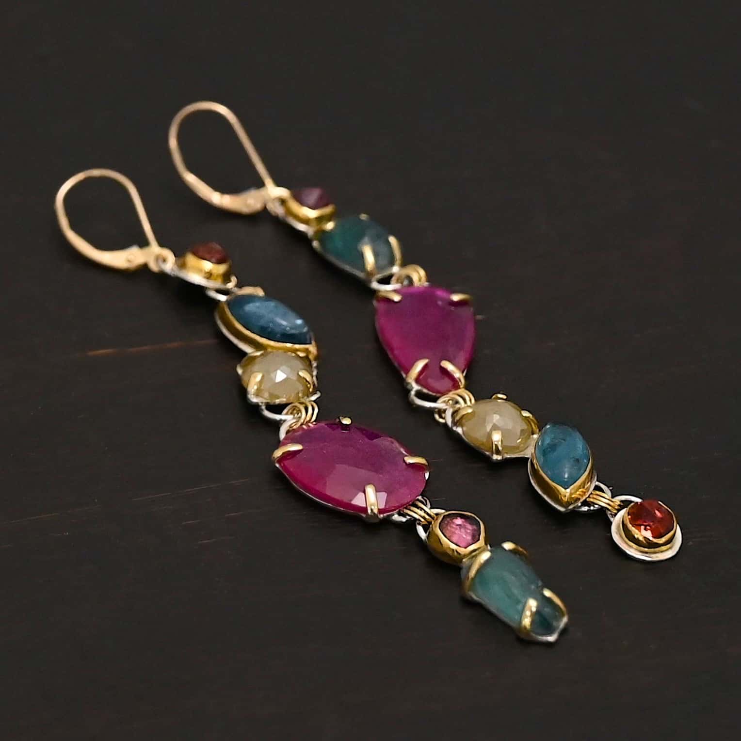 Follow the Path Earrings Photo of the "Follow Your Path" Earrings. Made out of sterling silver & 18K gold, tourmalines, apatites, rough diamonds, & rubies. They are approximately 3.5″ long. The rubies are in the center, and the rest of the stones are above or beneath them. The rubies are held in with gold prongs, as well as some of the other stones. Each stone setting has a loop on the back of it with gold jump rings that connect the stones to one another. Earrings have gold leverback ear wires. Earrings are laying against a dark background.