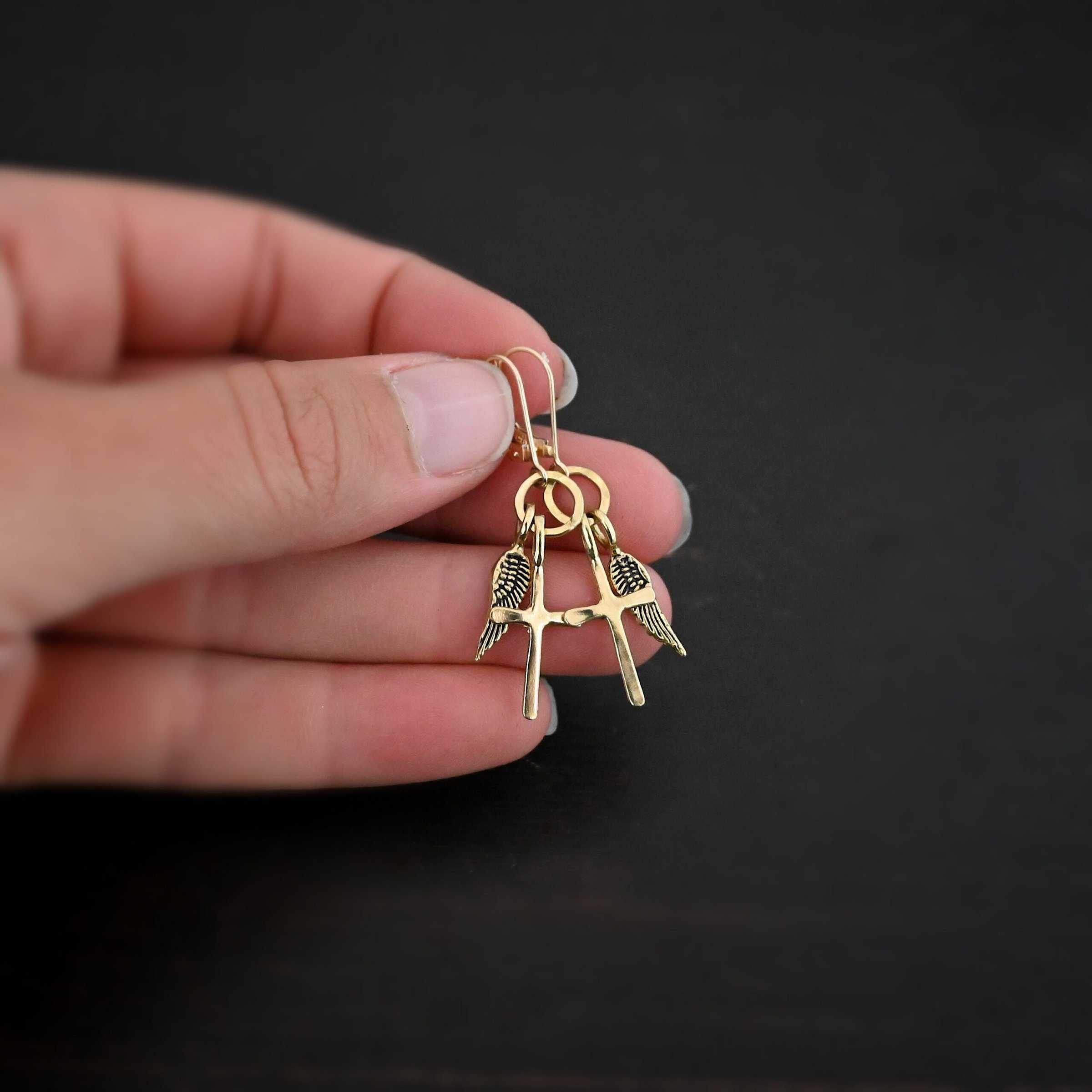 Strength Earrings Photo of the "Strength" Earrings. Made out of gold. There are a cross and angel wing charm on each earring. Both charms are small, and attached to a small hammered sterling silver loop that the French hook ear wires are attached to on the top. Earrings are held in the fingertips of a hand to demonstrate size.