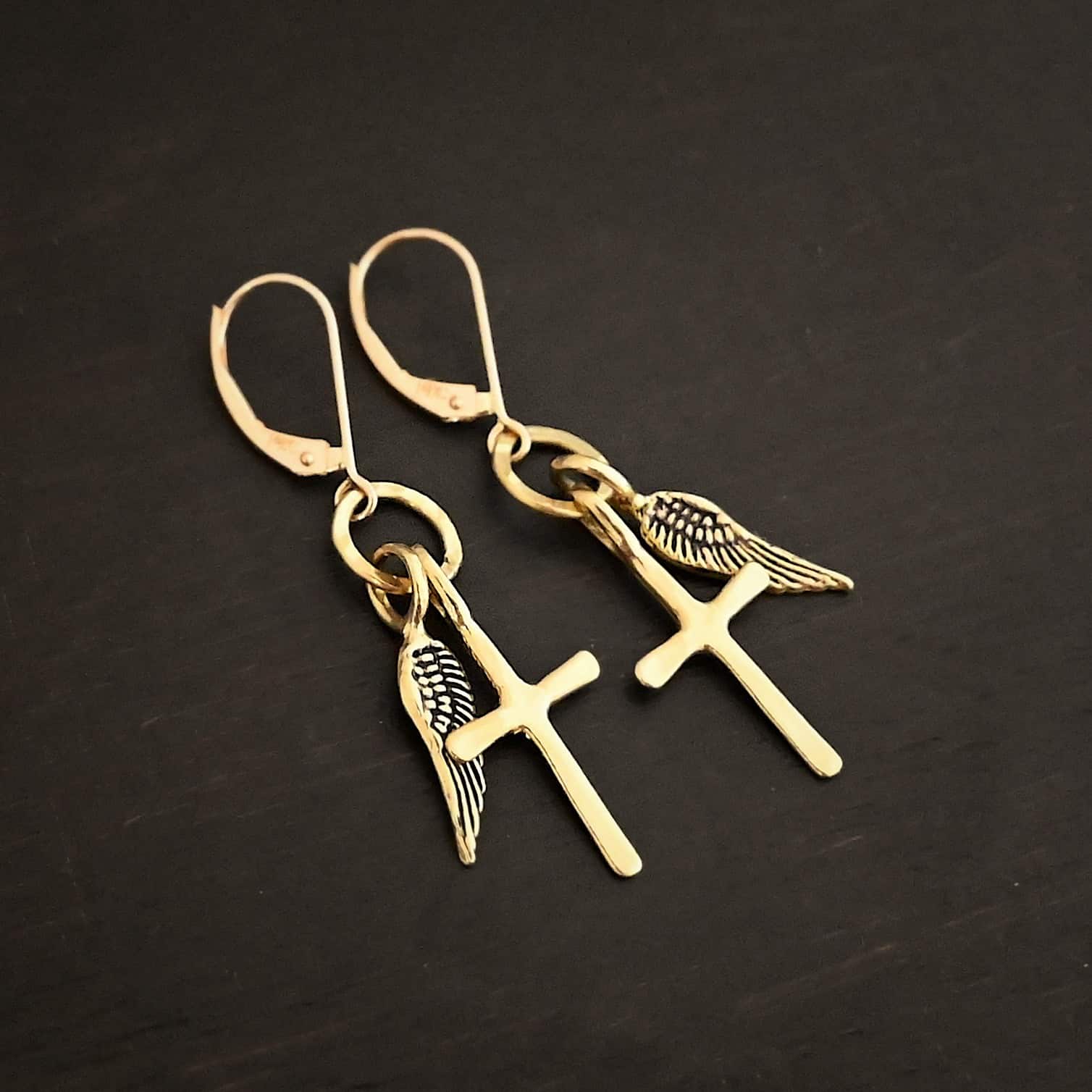 Strength Earrings Photo of the "Strength" Earrings. Made out of gold. There are a cross and angel wing charm on each earring. Both charms are small, and attached to a small hammered sterling silver loop that the French hook ear wires are attached to on the top. Earrings are shiny and laying against a dark background.