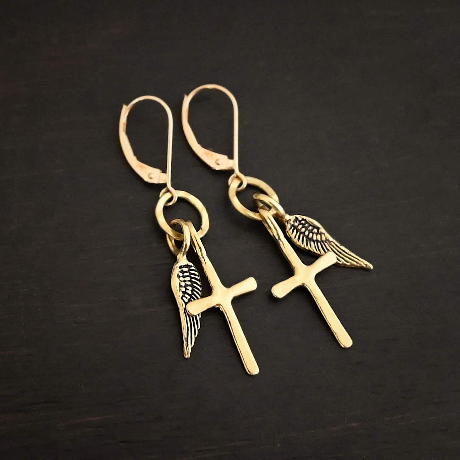 Strength Earrings Photo of the "Strength" Earrings. Made out of gold. There are a cross and angel wing charm on each earring. Both charms are small, and attached to a small hammered sterling silver loop that the French hook ear wires are attached to on the top. Earrings are shiny and laying against a dark background.