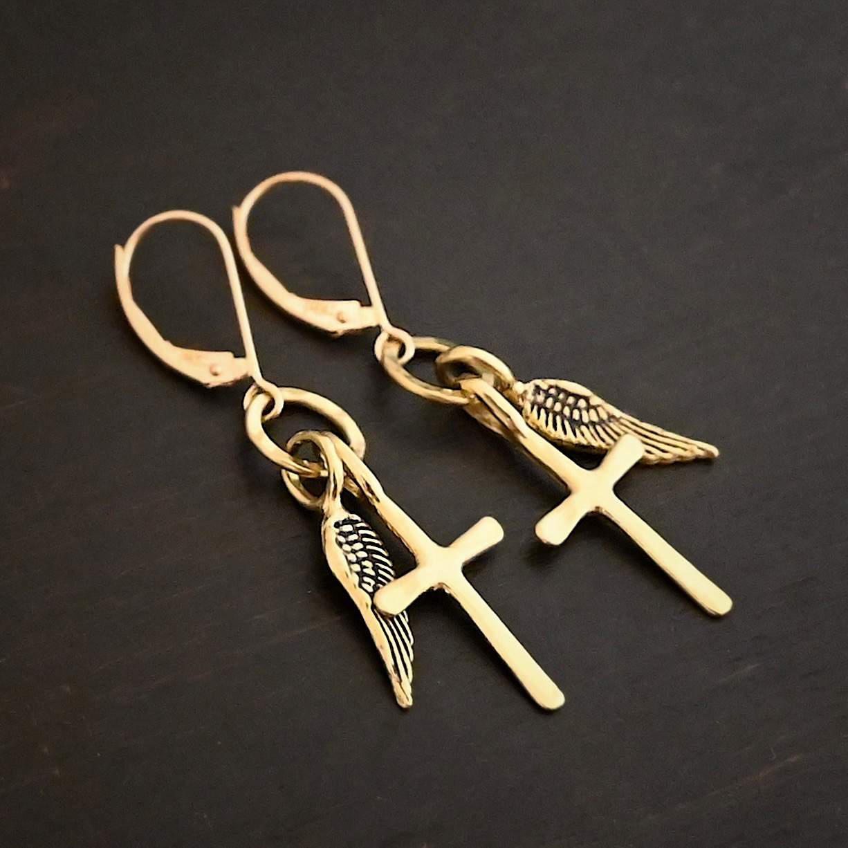 Strength Earrings Photo of the "Strength" Earrings. Made out of gold. There are a cross and angel wing charm on each earring. Both charms are small, and attached to a small hammered sterling silver loop that the French hook ear wires are attached to on the top. Earrings are shiny and laying against a dark background.
