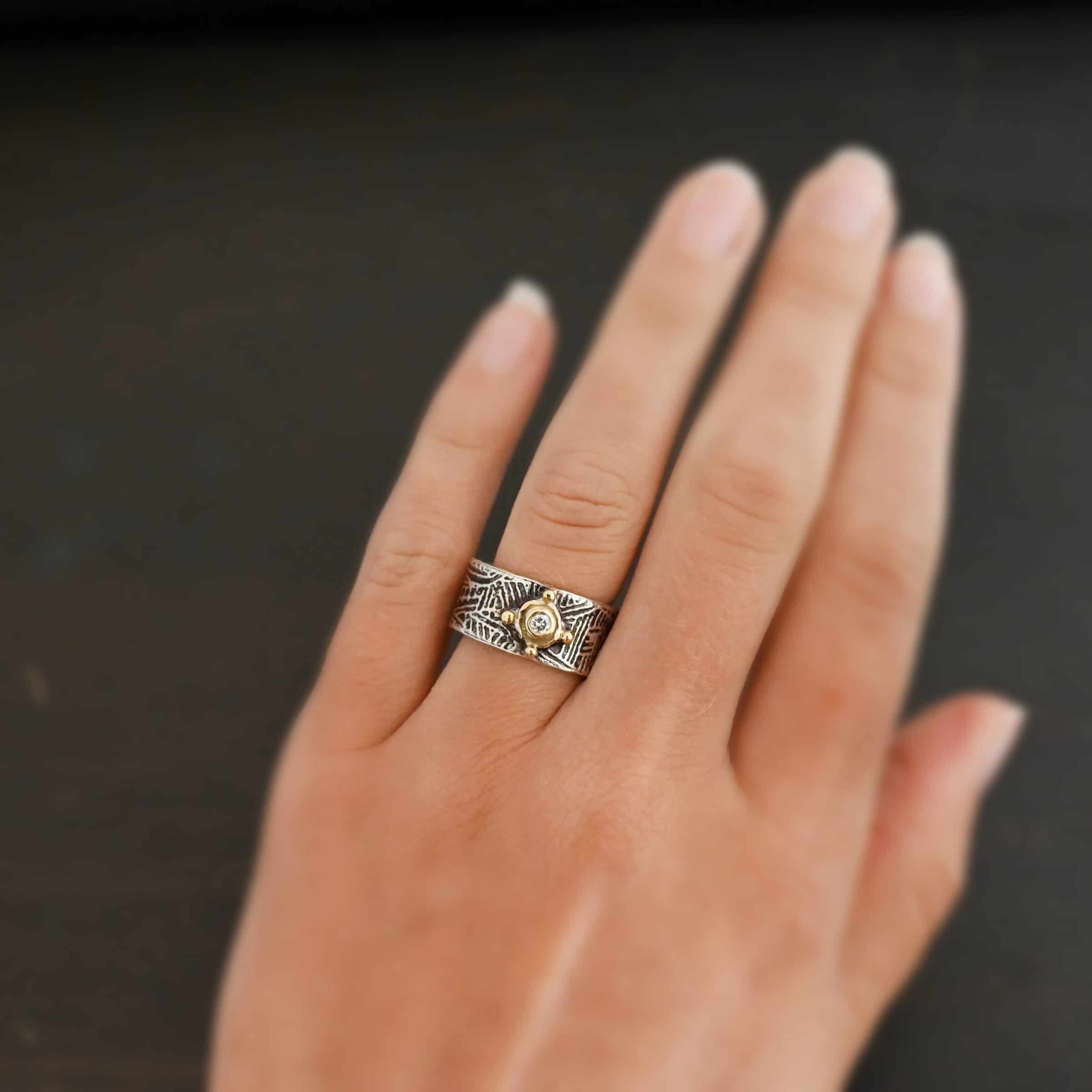 Castle Hill Ring Photo of the "Castle Hill" Ring. Made out of sterling silver, gold, and diamond. The center of the ring has a small diamond that is set in gold and sitting on a small gold dot. On each side of the big gold dot are four small gold dots. One is on the top, one on each side, and one on the bottom. The rest of the ring band is etched with lines that go every way and are different lengths. Ring is worn on a models engagement ring finger to demonstrate size.