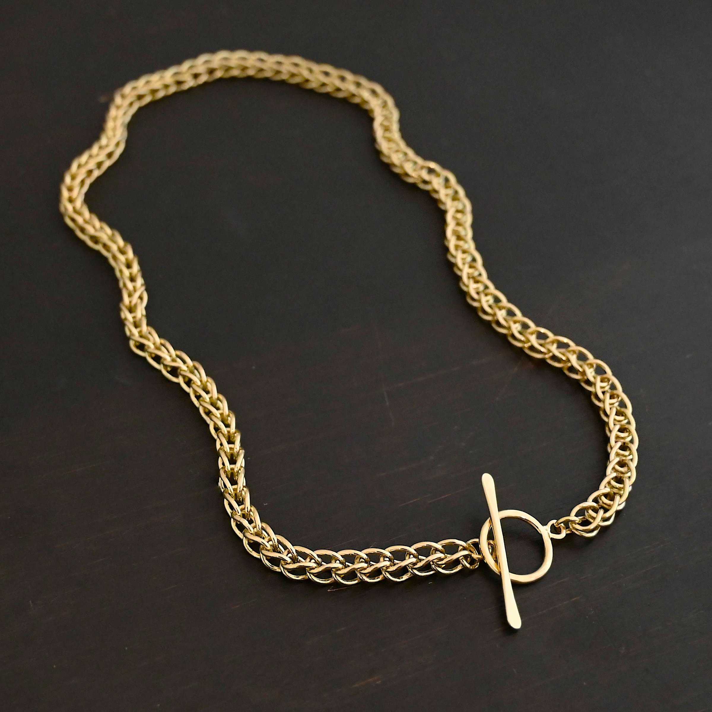 Gretchen Chain 18k Photo of a custom chain. The chain is solid gold, and thick. It is a viking knit chain with a large loop on the back with a toggle clasp. The chain is lying against a dark background.
