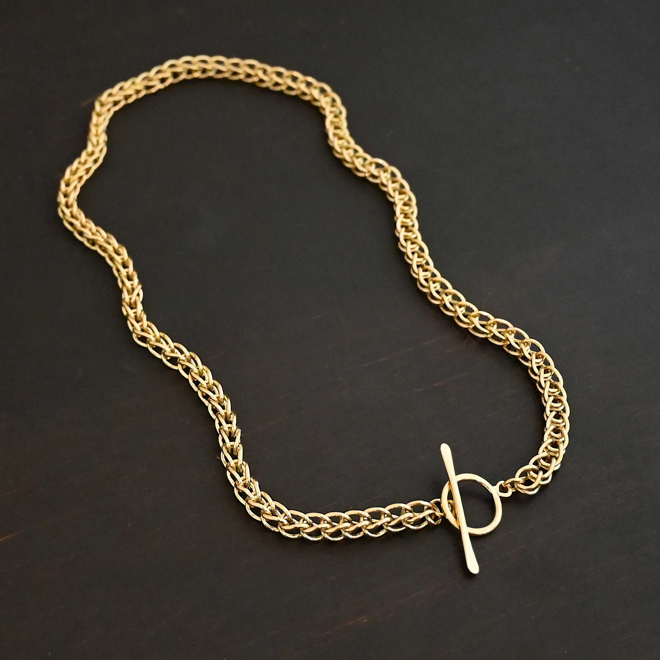 Gretchen Chain 18k Photo of a custom chain. The chain is solid gold, and thick. It is a viking knit chain with a large loop on the back with a toggle clasp. The chain is lying against a dark background.