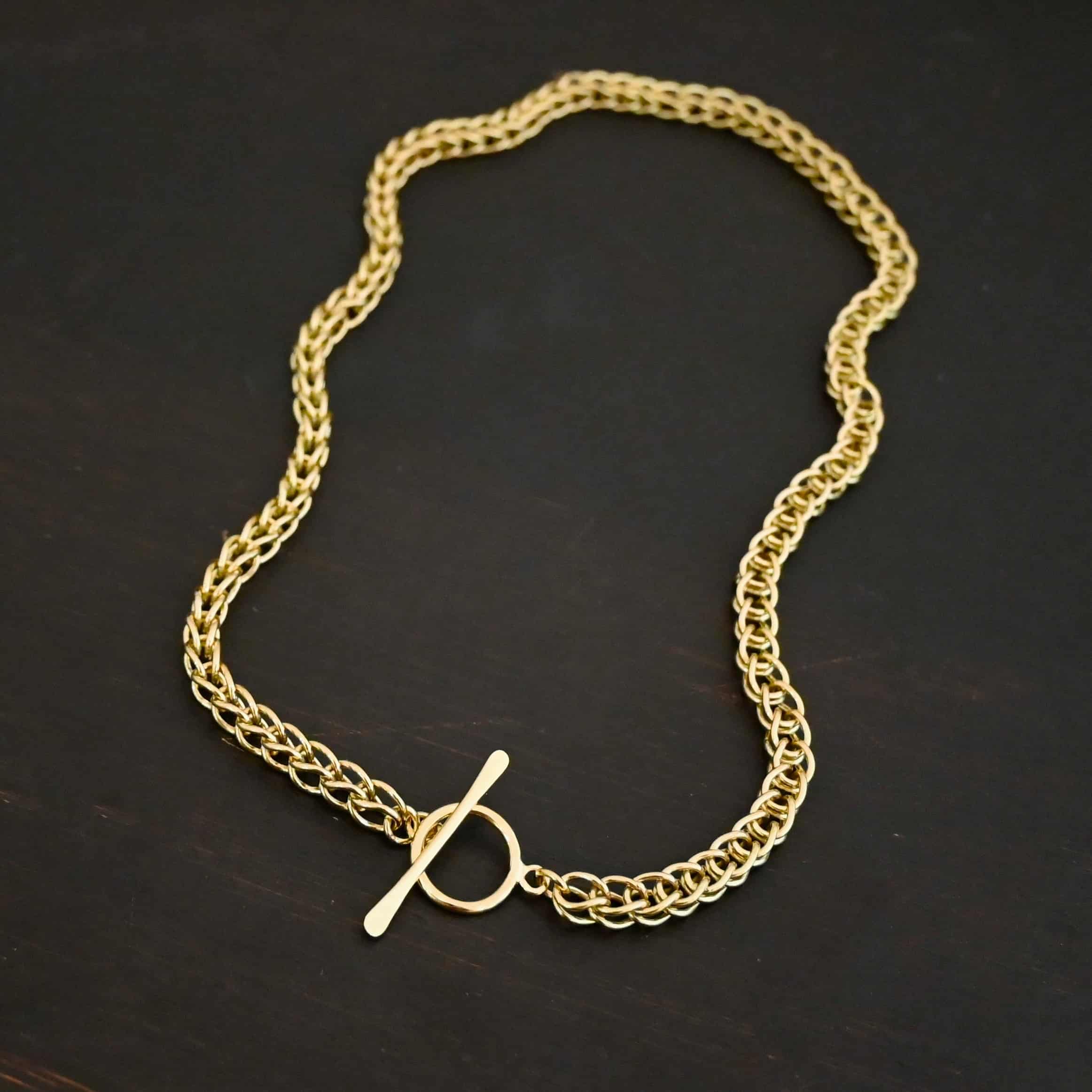 Gretchen Chain 18k Photo of a custom chain. The chain is solid gold, and thick. It is a viking knit chain with a large loop on the back with a toggle clasp. The chain is lying against a dark background.