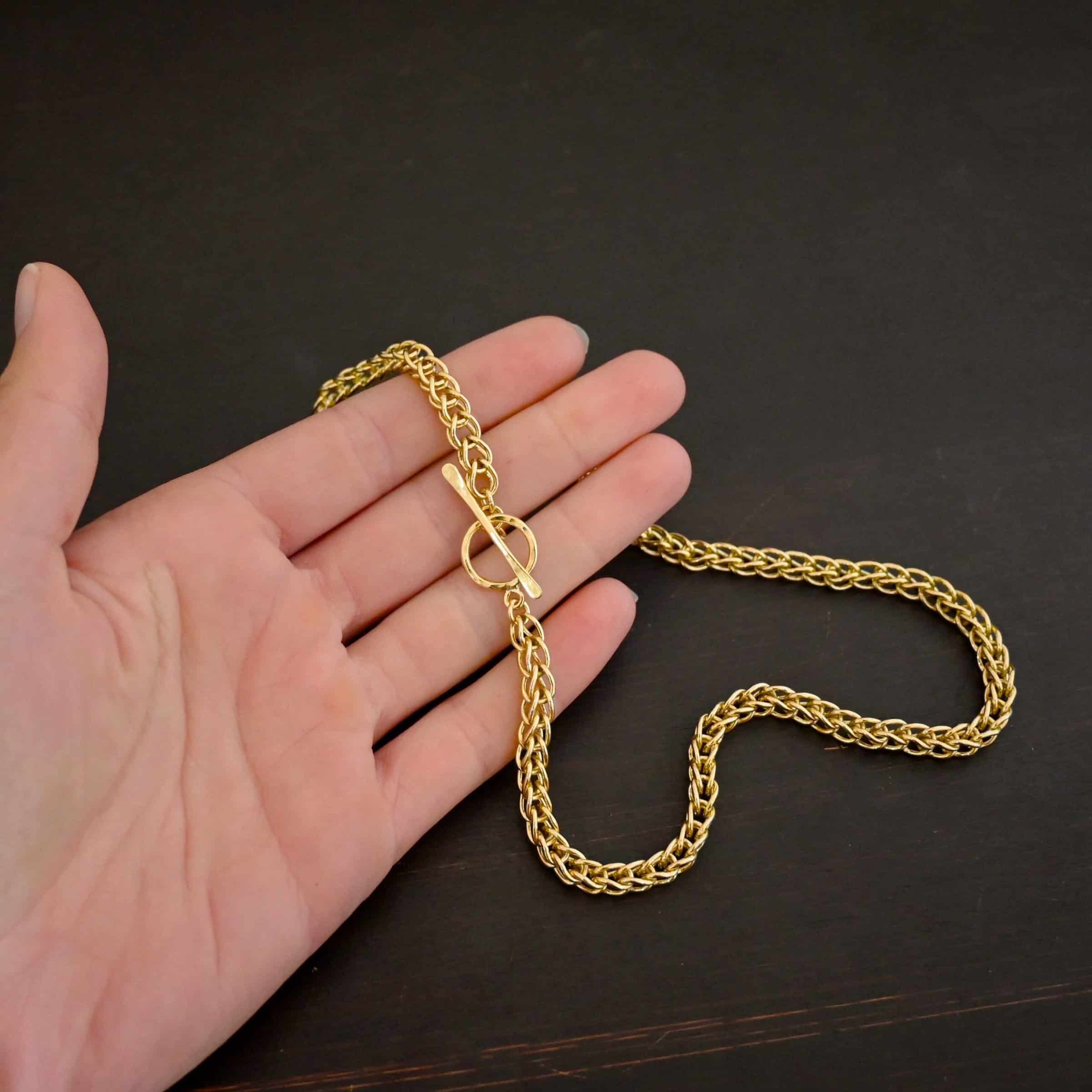 Gretchen Chain 18k Photo of a custom chain. The chain is solid gold, and thick. It is a viking knit chain with a large loop on the back with a toggle clasp. The chain is lying against the fingertips of a models hand to demonstrate size.