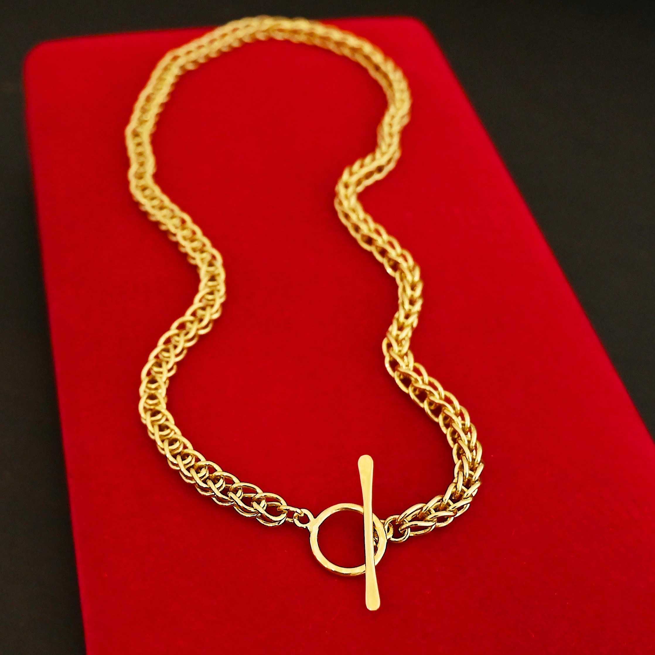 Gretchen Chain 18k Photo of a custom chain. The chain is solid gold, and thick. It is a viking knit chain with a large loop on the back with a toggle clasp. The chain is lying against a long bright red velvet box.