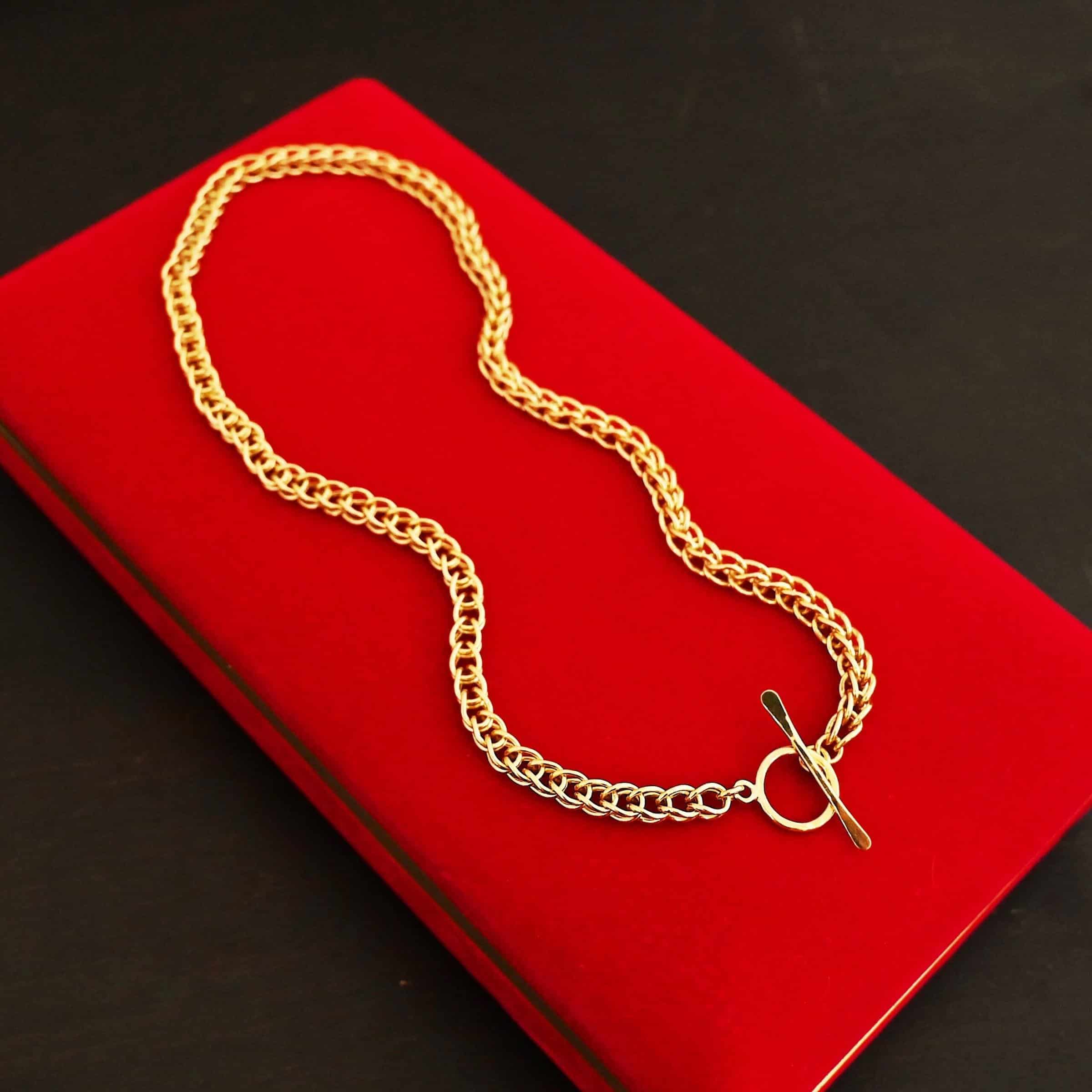 Gretchen Chain 18k Photo of a custom chain. The chain is solid gold, and thick. It is a viking knit chain with a large loop on the back with a toggle clasp. The chain is lying against a long bright red velvet box.