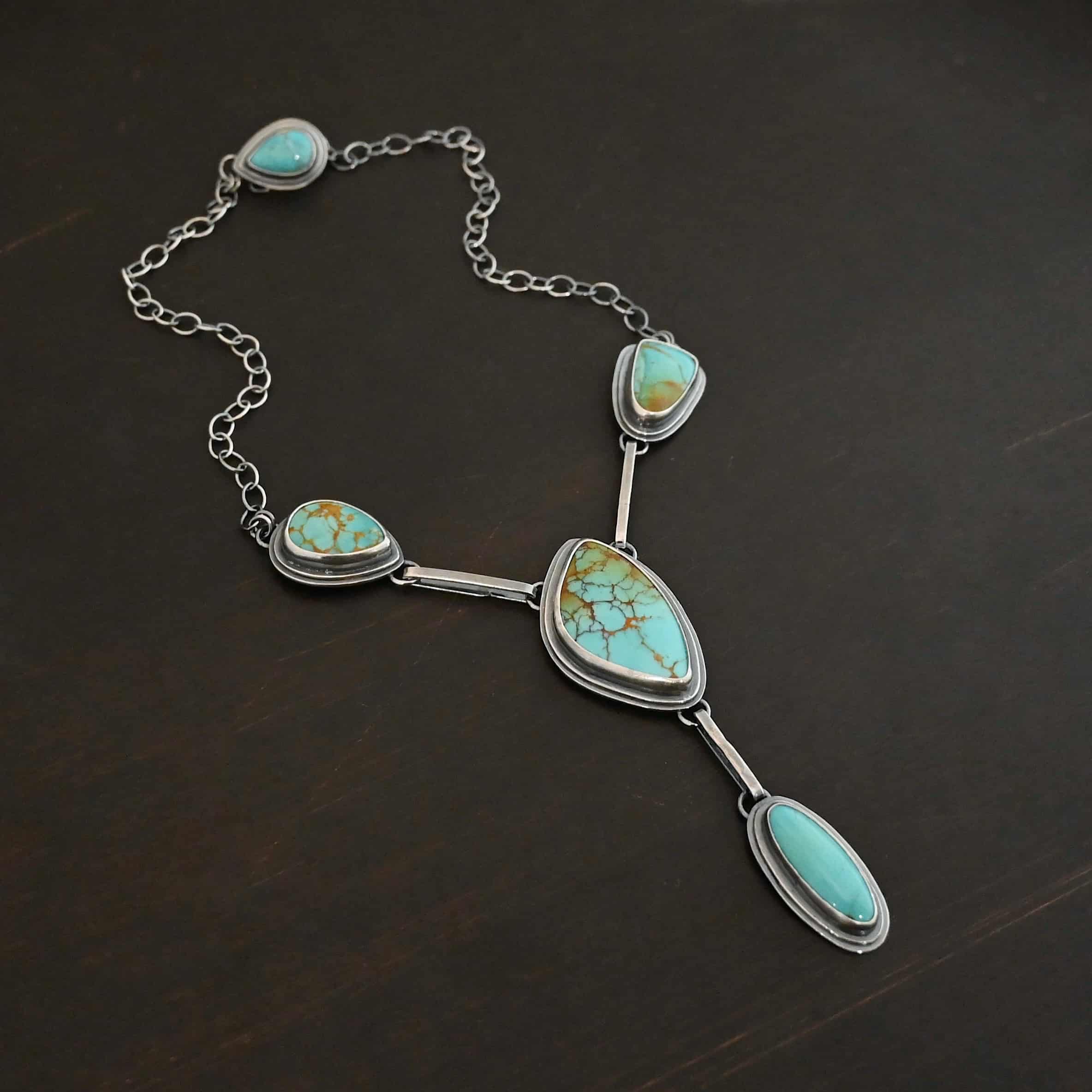 Gone with the Wind Necklace Photo of "Gone With The Wind" Necklace. This is a large turquoise necklace. The necklace has five turquoise, one on the left side, one on the right side, and two in the center. The two in the center are one on top of the other. There is a turquoise on the clasp of the necklace as well. Each turquoise is connected by a small silver bar and each bar is connected to loops on the backs of the settings for the turquoise. Necklace is lying against a dark background.