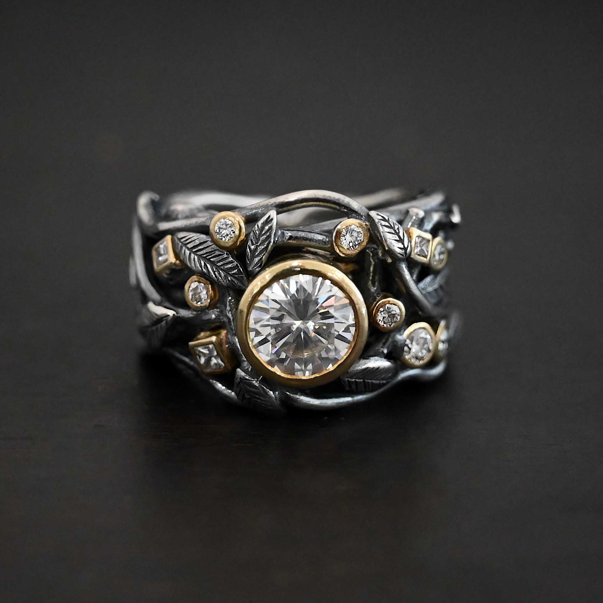 Enchanted Ring Photo of the "Enchanted" Ring. Ring has a large moissanite in the center and smaller moissanites scattered around the entire ring. There are multiple silver leaves around the entire ring. There are eleven stones total, the center stone, and ten moissanites around the rest of the ring, five on each side. Each stone is set in gold. Ring is set against a dark background.