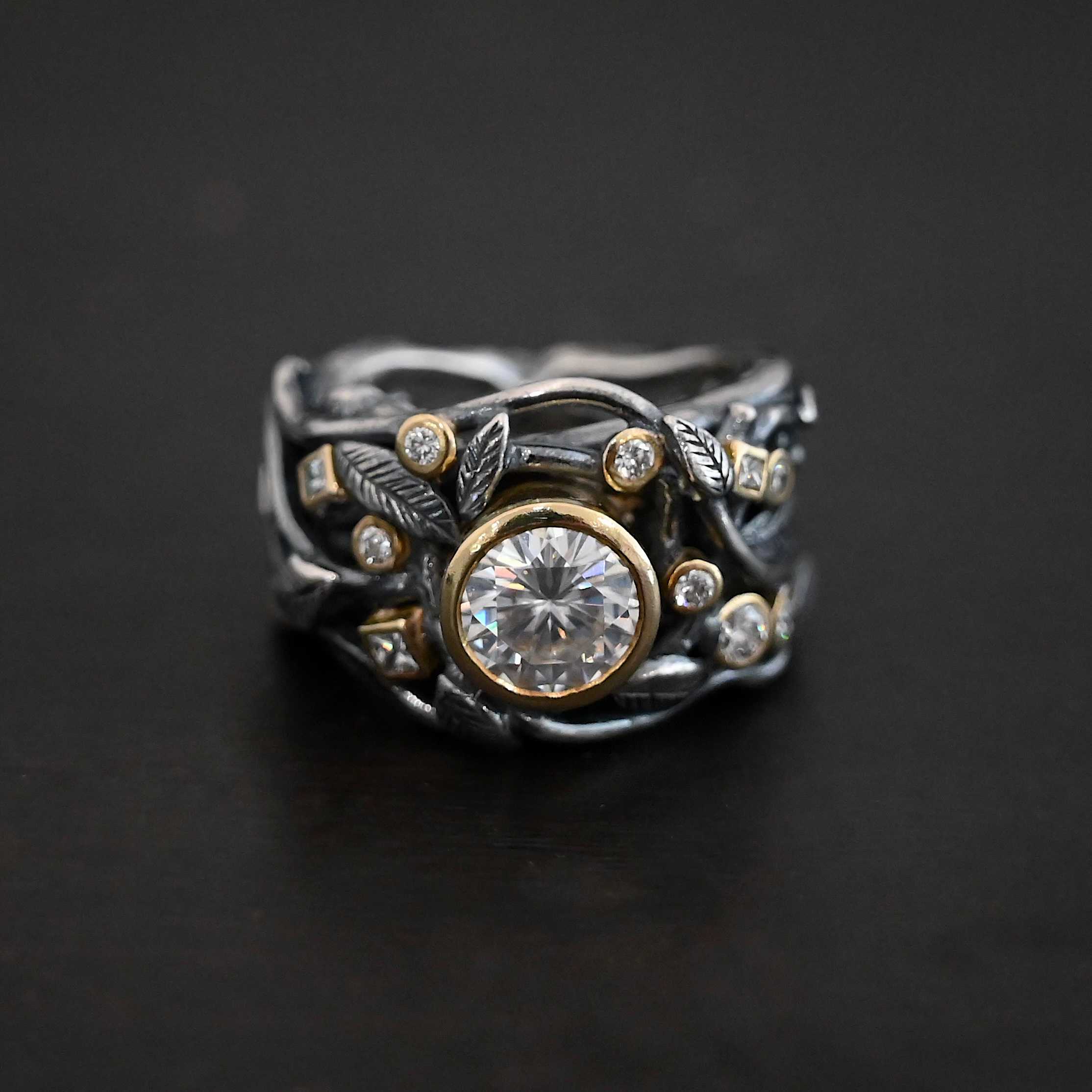 Enchanted Ring Photo of the "Enchanted" Ring. Ring has a large moissanite in the center and smaller moissanites scattered around the entire ring. There are multiple silver leaves around the entire ring. There are eleven stones total, the center stone, and ten moissanites around the rest of the ring, five on each side. Each stone is set in gold. Ring is set against a dark background.