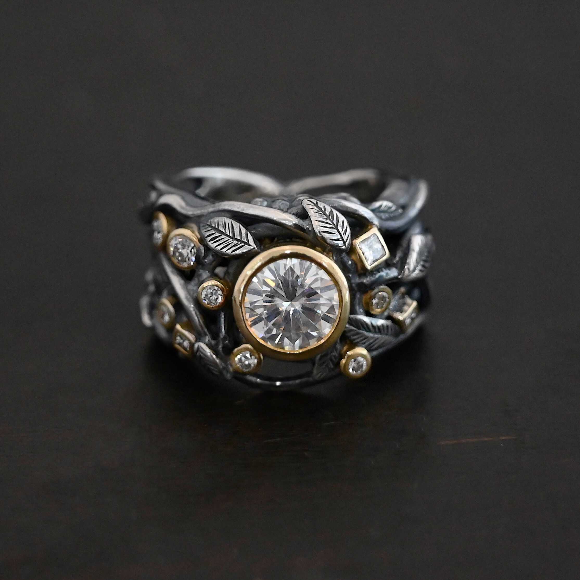 Enchanted Ring Photo of the "Enchanted" Ring. Ring has a large moissanite in the center and smaller moissanites scattered around the entire ring. There are multiple silver leaves around the entire ring. There are eleven stones total, the center stone, and ten moissanites around the rest of the ring, five on each side. Each stone is set in gold. Ring is set against a dark background.